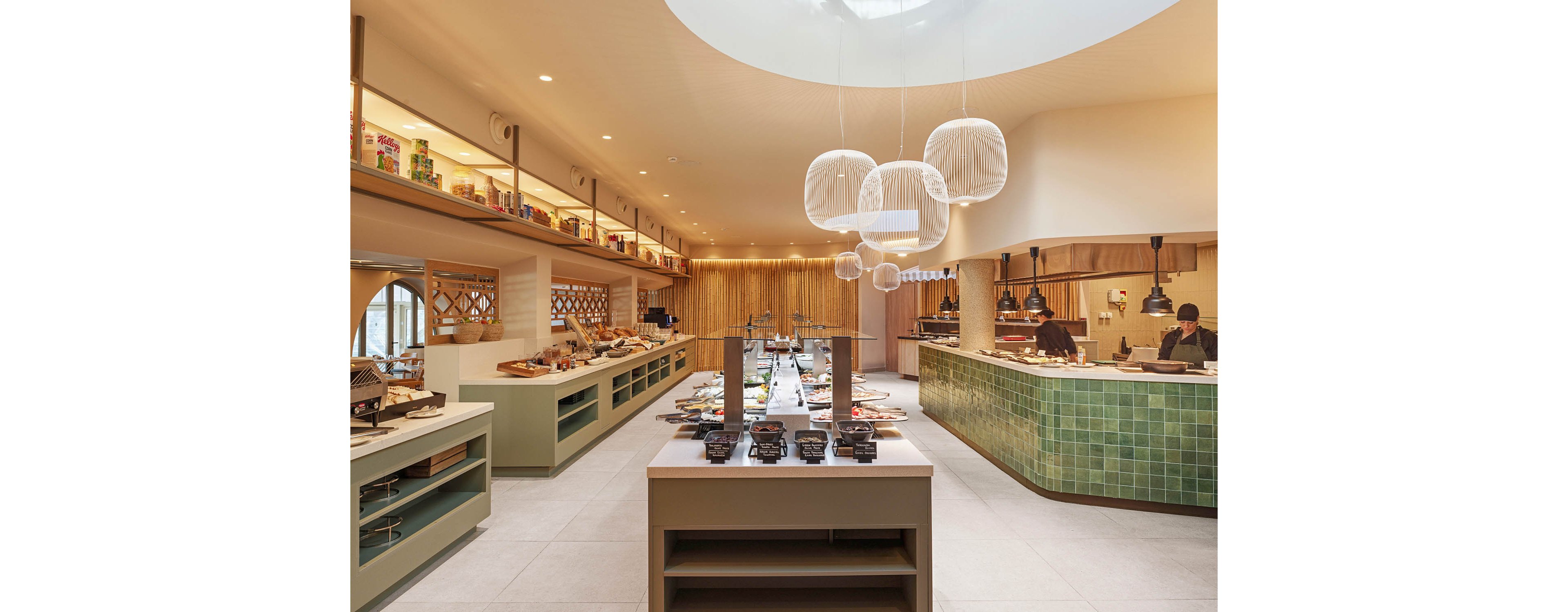 A contemporary buffet restaurant interior features long counters filled with assorted dishes and condiments. The space includes an open kitchen area where staff prepare food behind a green-tiled counter. Warm wooden accents, soft beige tones, and hanging woven light fixtures create a welcoming atmosphere. A large circular skylight brightens the room, highlighting the clean, minimalist design. The overall setting suggests a self-service dining experience in a stylish, modern venue.