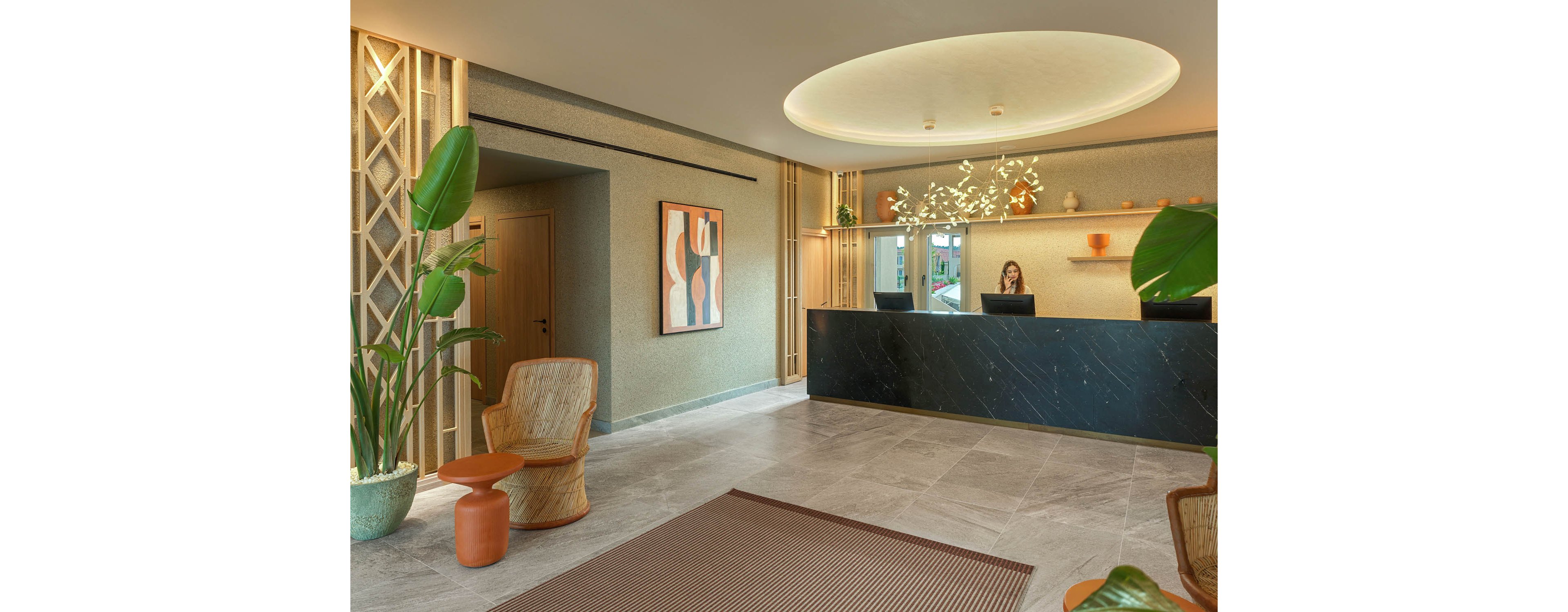 A contemporary hotel reception area features a sleek black front desk where a receptionist works at a computer. The space is decorated with large potted plants, woven chairs, and a small side table, creating a welcoming lounge corner. Soft neutral tones, wood accents, and an abstract wall painting enhance the modern aesthetic. A circular ceiling light and decorative hanging foliage add warmth and sophistication to the lobby.