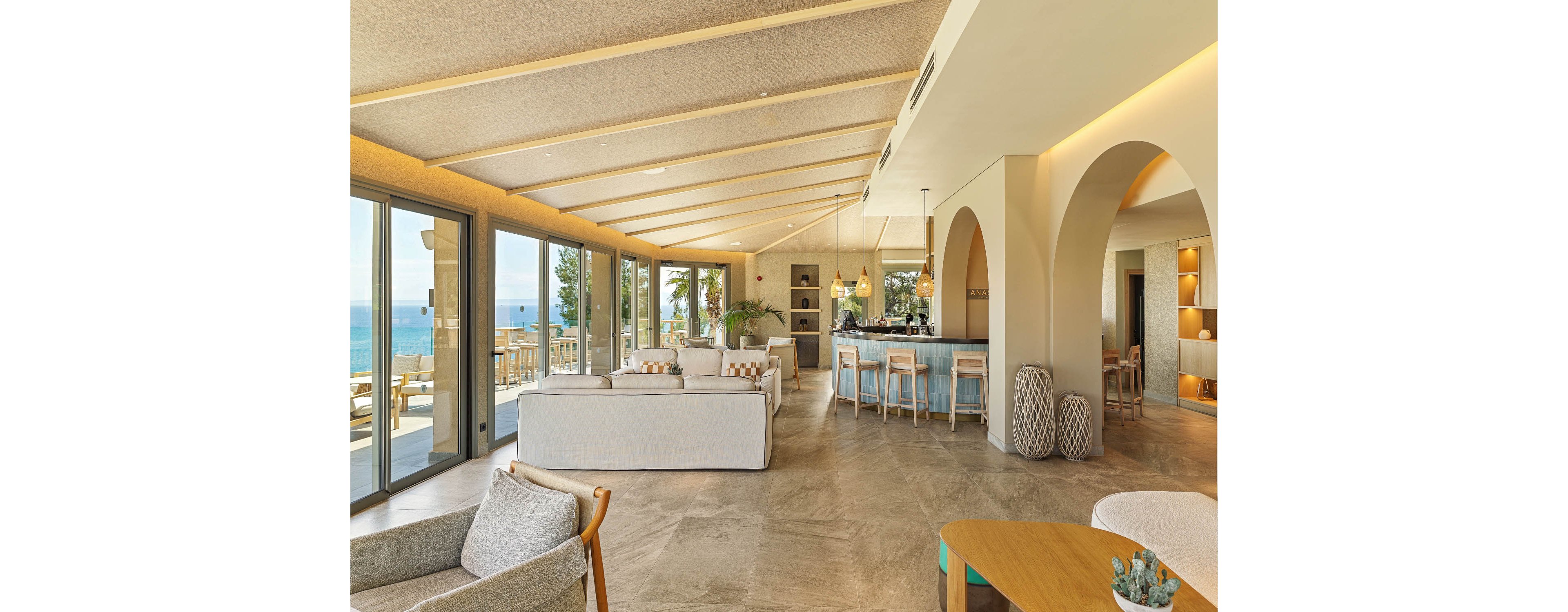A spacious modern lounge features neutral-toned sofas and armchairs arranged around a central seating area. Large floor-to-ceiling windows open to a terrace with views of the ocean and palm trees. On the right, a contemporary bar with high stools sits beneath arched openings and pendant lights. Warm beige and wood finishes create a relaxed, coastal atmosphere with abundant natural light.