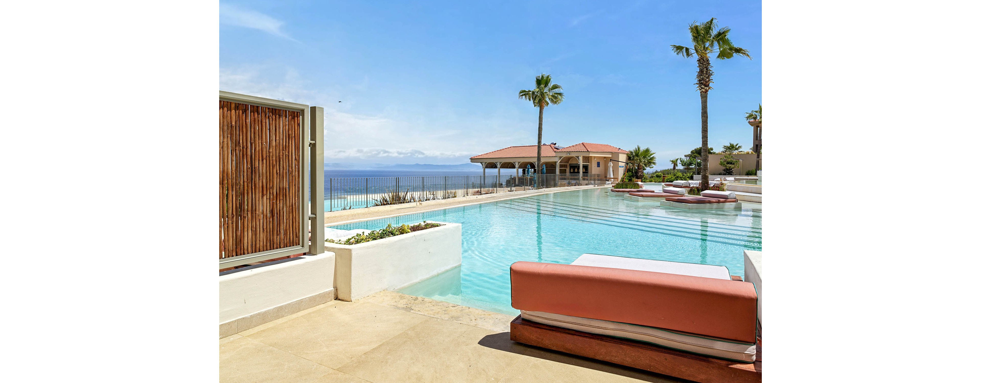 A modern infinity pool stretches toward a distant ocean horizon at a coastal resort. Pink cushioned loungers and built-in seating areas line the pool, creating a relaxed and upscale atmosphere. Palm trees rise above the water, and a light-colored pool deck leads toward an open-air pavilion. Clear blue skies and calm water emphasize a serene, sunny vacation setting.