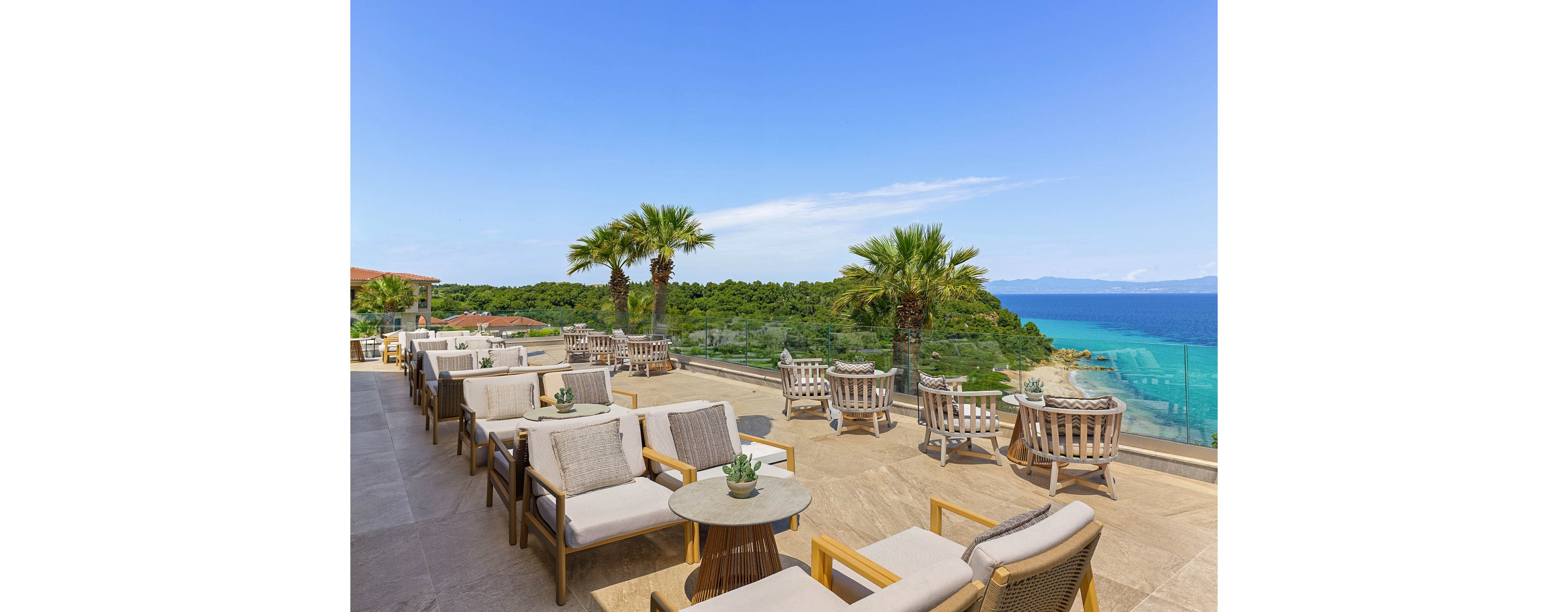An elevated outdoor terrace lounge overlooks a bright blue coastline and lush green hills. Comfortable cushioned chairs and small round tables are arranged in rows, creating a relaxed seating area. Palm trees line the glass railing, framing the expansive sea and clear sky. The scene suggests a tranquil resort or hotel setting ideal for relaxation and social gatherings.