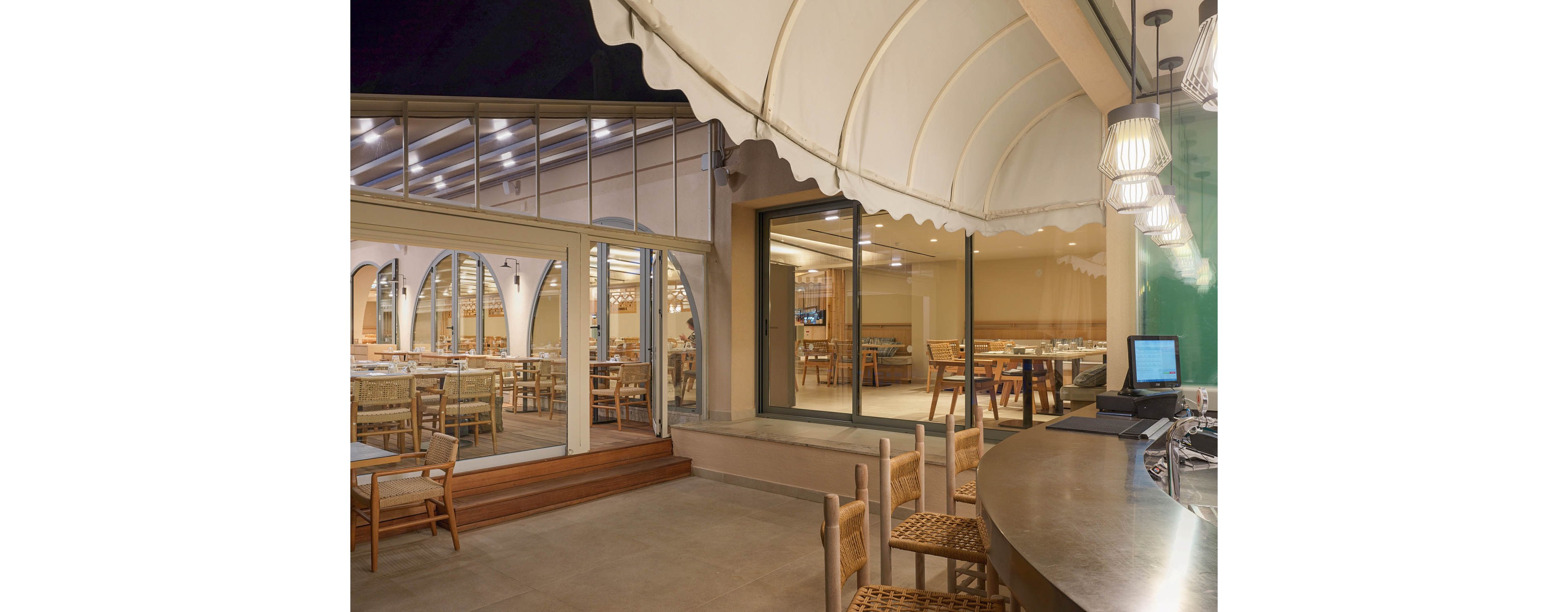A contemporary restaurant terrace is shown at night with warm interior lighting visible through large glass windows. Wooden chairs and tables are arranged both inside and outside, creating an inviting dining atmosphere. A curved bar counter with pendant lights and a computer screen sits in the foreground. The arched window reflections and fabric canopy overhead add architectural interest and a cozy, sheltered feel.