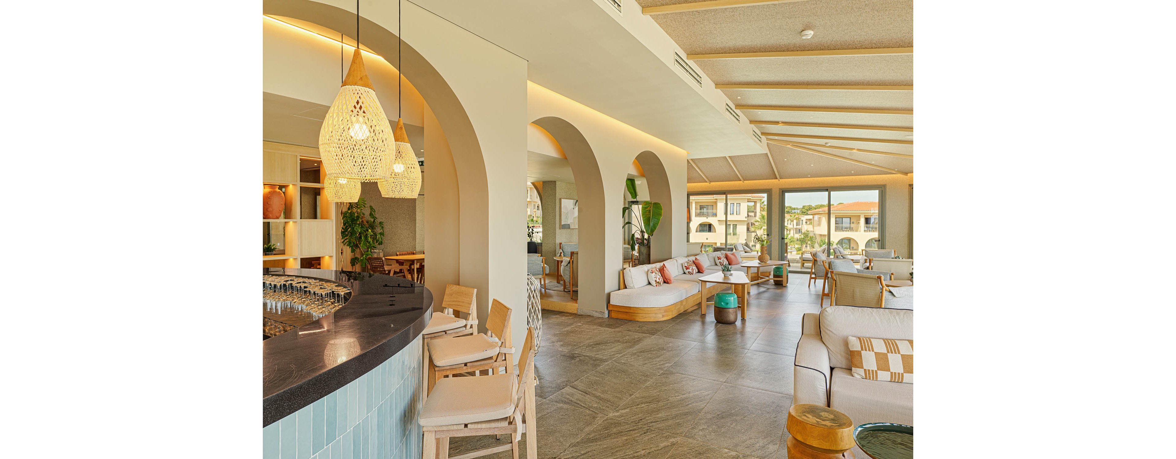 Spacious hotel lounge with a curved bar and modern seating is bathed in warm natural light. Large arched openings and floor-to-ceiling windows reveal a resort-style courtyard outside. Neutral tones, woven pendant lights, and wooden accents create a relaxed Mediterranean atmosphere. Cushioned sofas, stools, and plants add comfort and a casual social vibe to the contemporary space.