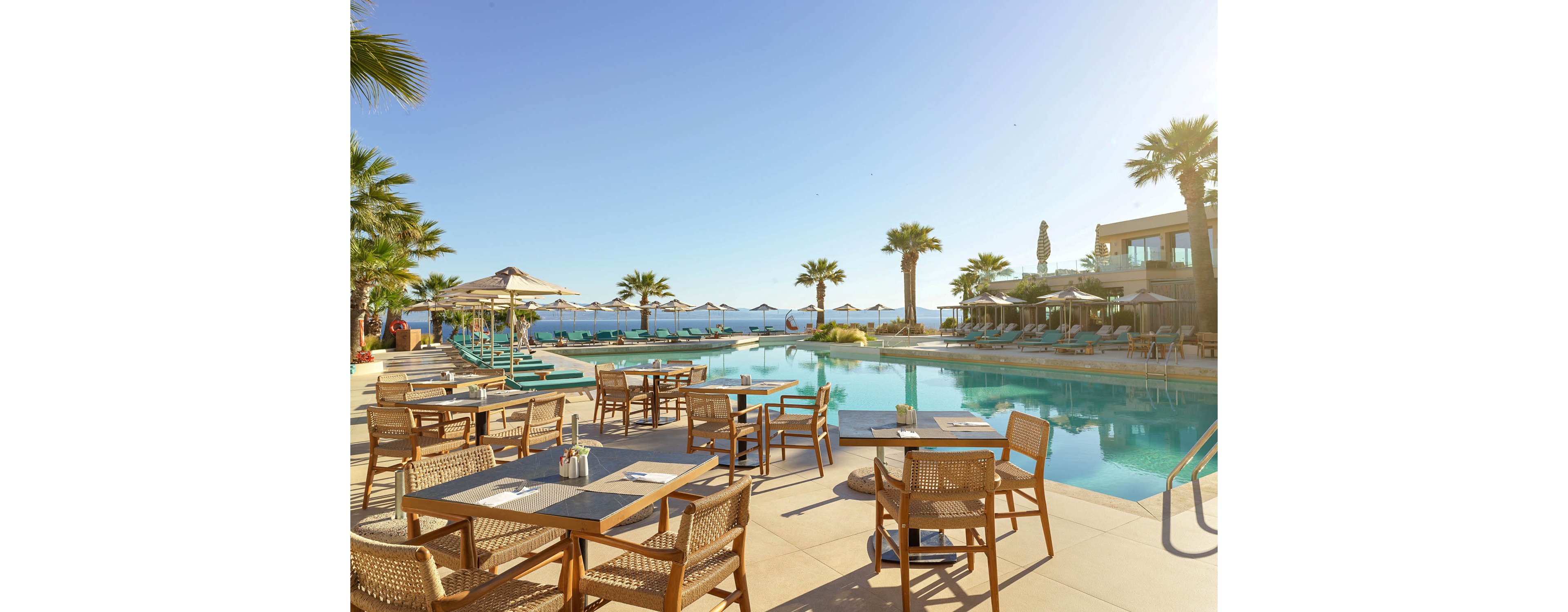 Outdoor restaurant tables overlook a large swimming pool at a seaside resort. Sun loungers and umbrellas line the pool, with calm blue water and the ocean visible in the background. Palm trees frame the scene under a clear sky, creating a relaxed vacation atmosphere. The warm sunlight enhances the tranquil, luxurious setting.