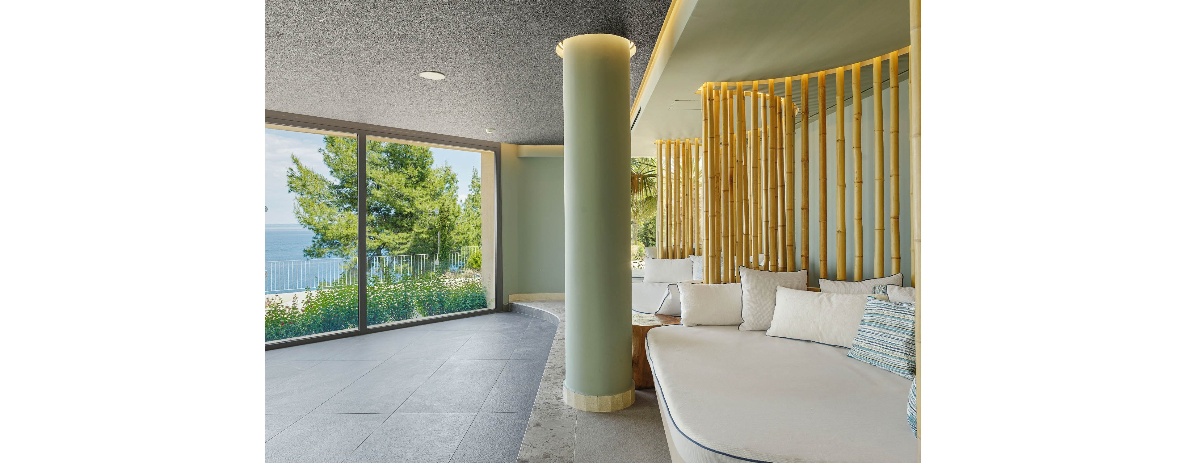 Contemporary indoor lounge area features a curved built-in sofa with light cushions and a cylindrical column in the center. Floor-to-ceiling windows open to a scenic view of trees, railing, and the distant sea. Vertical bamboo-style slats add a natural accent to the soft green walls and minimalist decor. The space feels calm and airy, combining modern architecture with a serene coastal landscape.