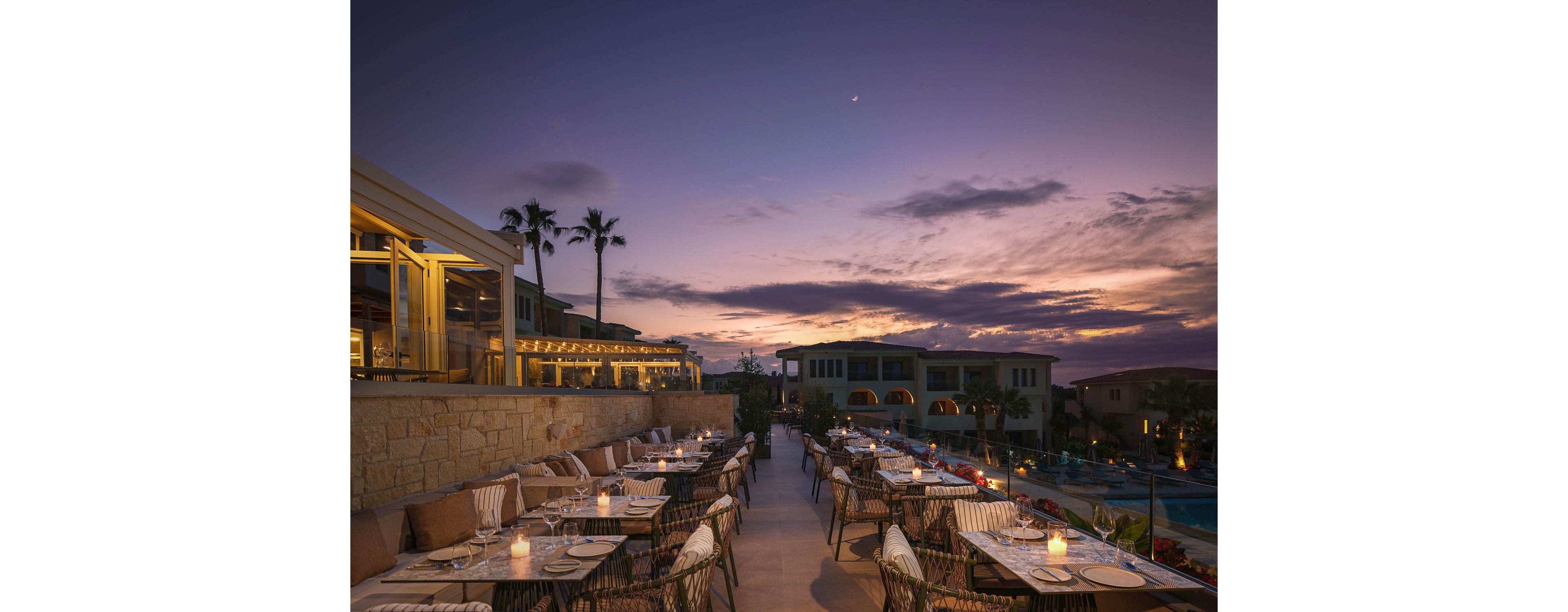 An elegant rooftop restaurant is set for evening dining under a dramatic sunset sky. Candlelit tables with neatly arranged place settings line a terrace overlooking a resort pool and surrounding buildings. Warm interior lights from the adjacent structure contrast with the deep blue and purple tones of the dusk sky. A few palm trees frame the scene, enhancing the relaxed, upscale vacation atmosphere.