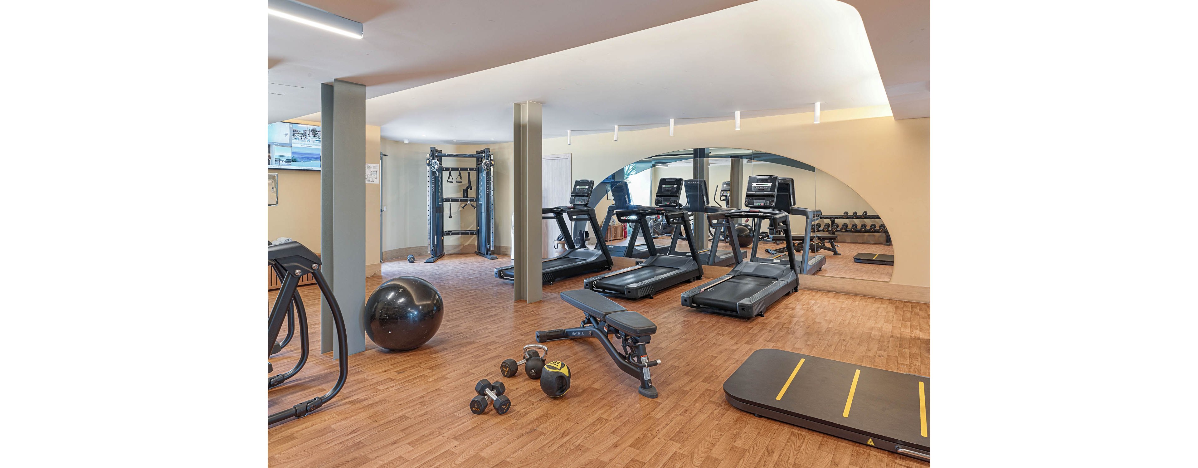 A bright, modern gym features several treadmills lined up against a mirrored wall. In the foreground, free weights, a weight bench, and a large exercise ball are arranged on a wooden floor. A cable machine stands to the side, offering additional strength training options. The neutral walls and clean lighting create a calm, inviting workout environment.