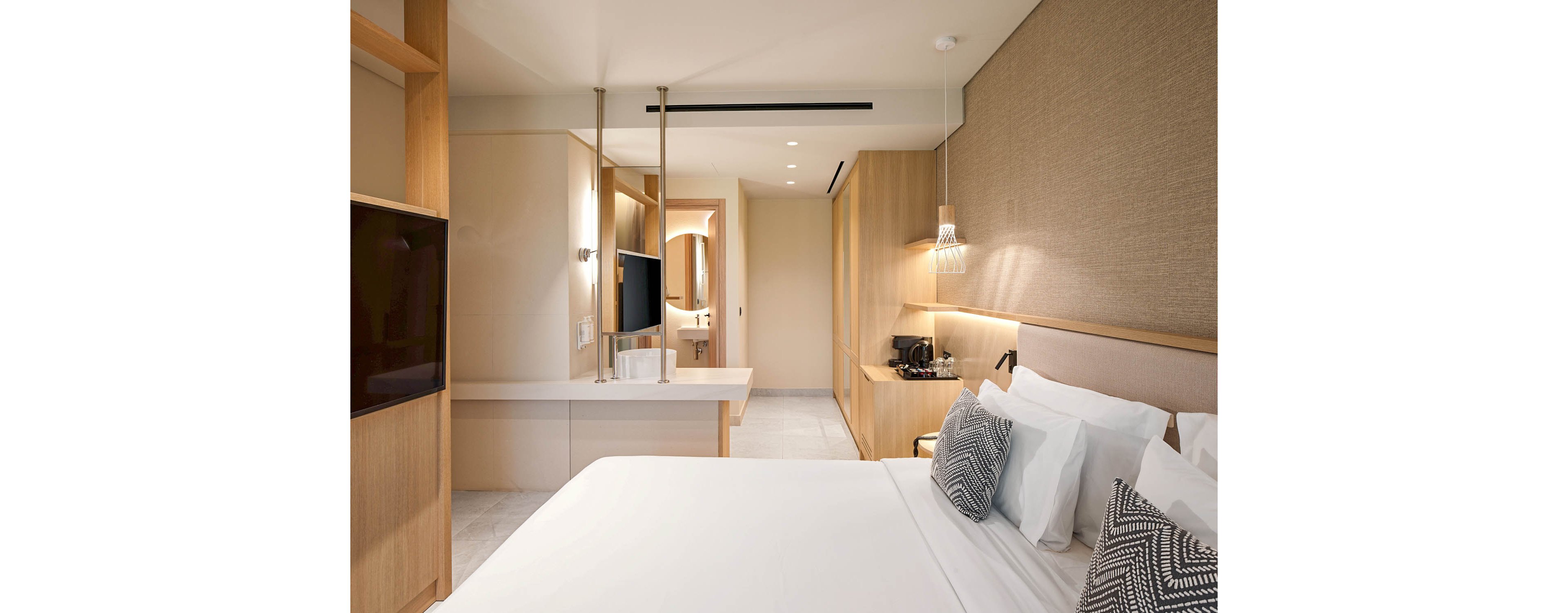 A contemporary hotel room features a neatly made bed with white linens and patterned accent pillows. Light wood furniture and paneling create a warm, minimalist atmosphere, complemented by a wall-mounted television and built-in desk area. Soft lighting, a hanging bedside lamp, and a coffee station add comfort and functionality. The open layout leads toward a bright bathroom area, enhancing the spacious feel of the room.