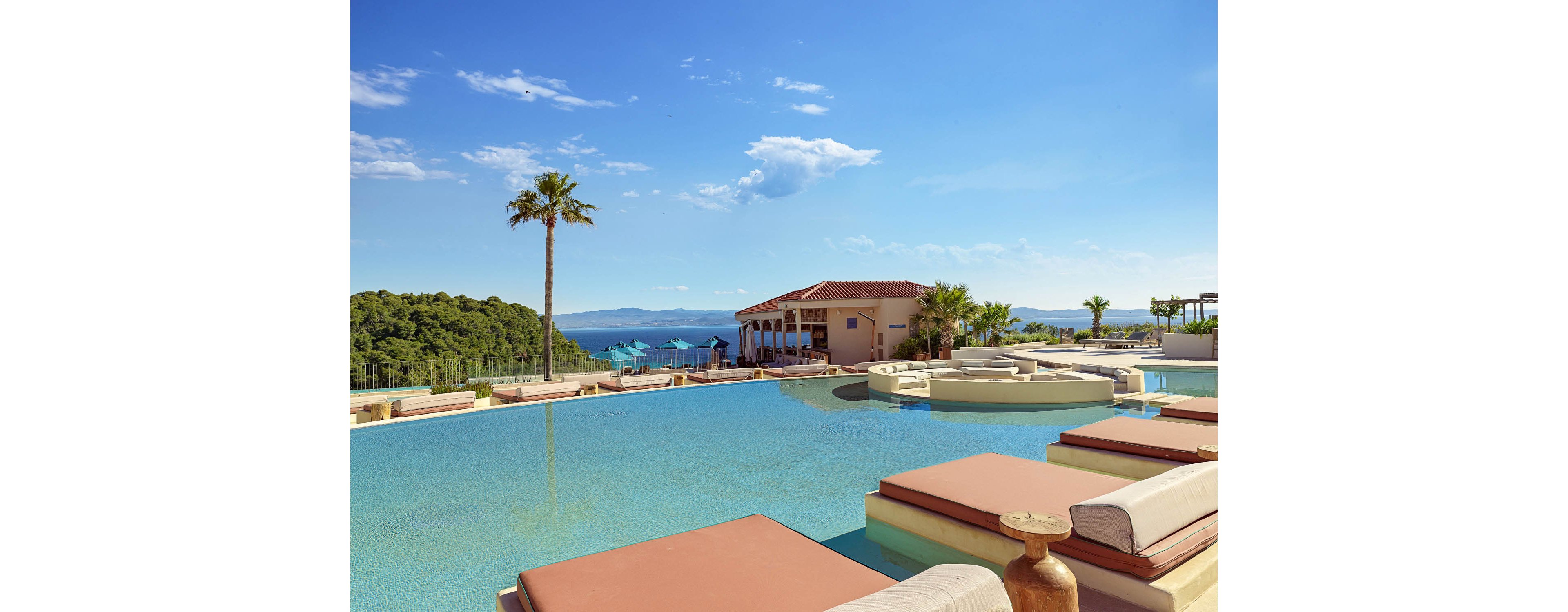 A luxurious infinity pool stretches toward a scenic coastal horizon with calm blue water. Cushioned sunbeds and circular lounge platforms surround the pool, suggesting a relaxing resort atmosphere. A low building with a red-tiled roof sits near the water, framed by palm trees and lush green hills. Clear skies and distant mountains enhance the tranquil, upscale vacation setting.