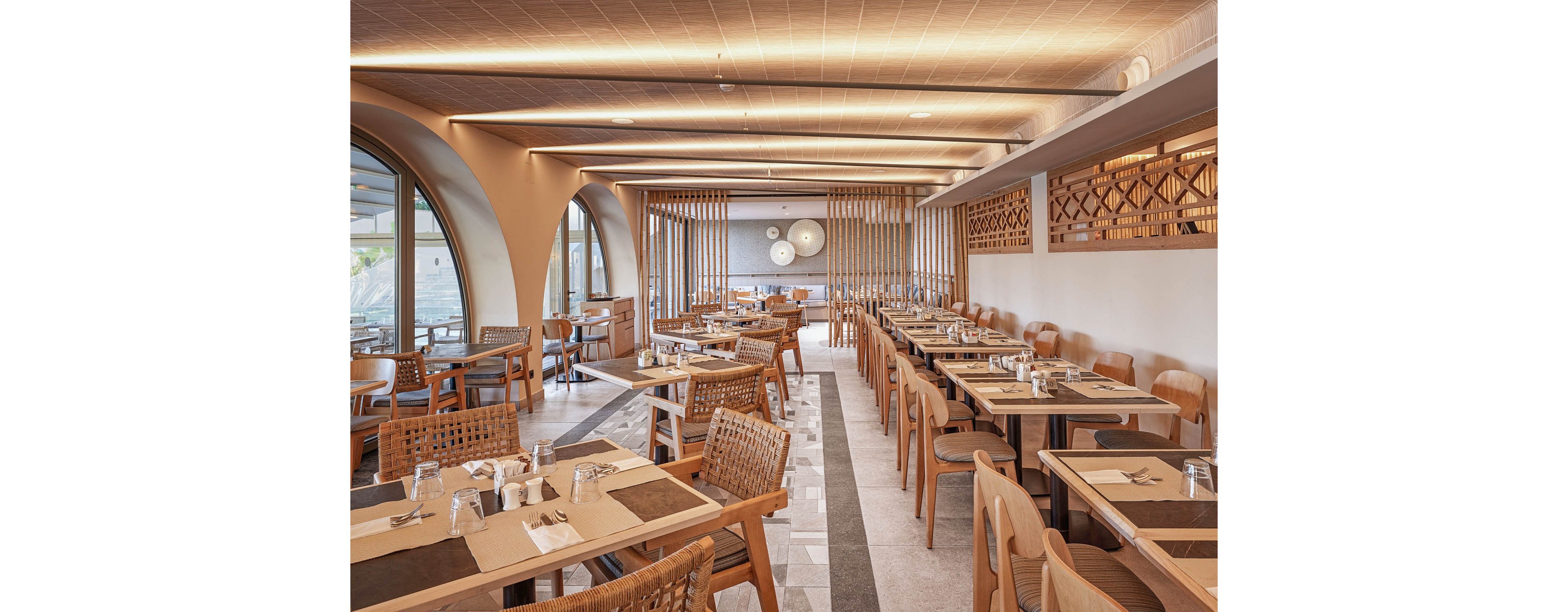 A contemporary restaurant dining area features neatly arranged wooden tables and woven chairs. The space is empty, with place settings of glasses, napkins, and cutlery prepared for guests. Warm lighting, arched windows, and decorative wooden partitions create a cozy yet modern atmosphere. Neutral tones and clean lines emphasize a minimalist, sophisticated design suitable for upscale dining.
