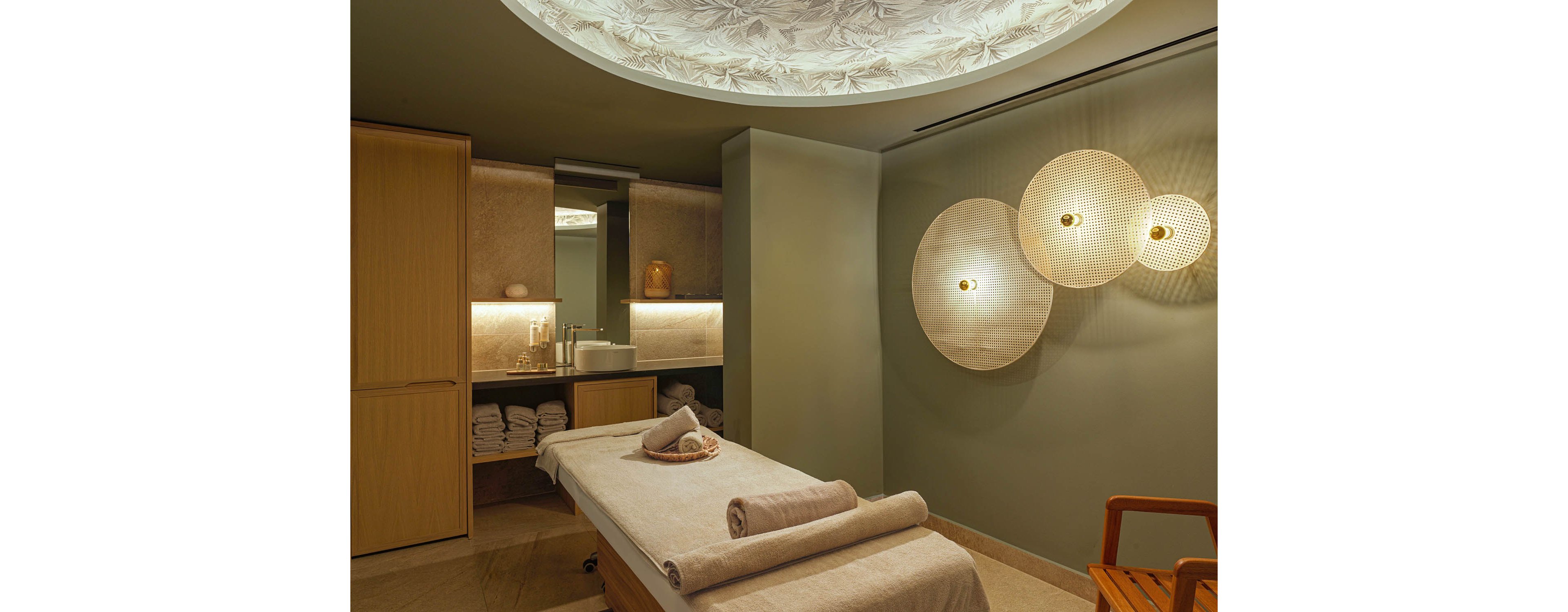 A tranquil spa treatment room features a massage table neatly prepared with rolled towels and a small decorative cushion. Warm, ambient lighting illuminates the space, highlighting textured wall panels and circular woven wall lights. Built-in cabinetry and open shelves hold neatly stacked towels and spa essentials near a modern sink area. The overall atmosphere is calm and inviting, with earthy tones and a soothing, contemporary design.