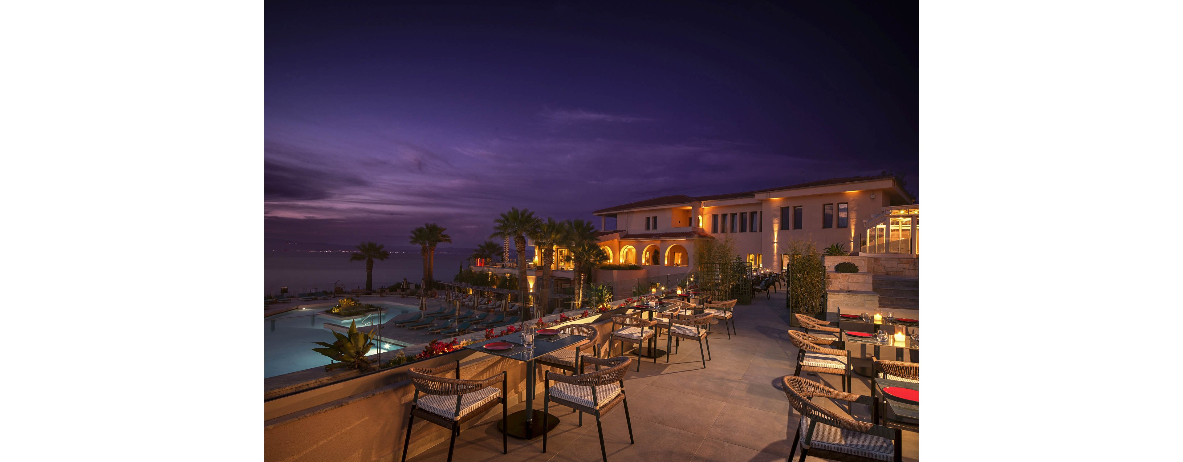 An elegant seaside resort terrace is set for evening dining, overlooking a lit swimming pool and calm water beyond. Tables and chairs are neatly arranged along the stone patio, with candles and soft lighting creating a romantic atmosphere. The resort building glows warmly with arched windows and balconies, contrasting against the deep purple twilight sky. Palm trees and landscaped greenery frame the scene, enhancing the tranquil, upscale ambiance.