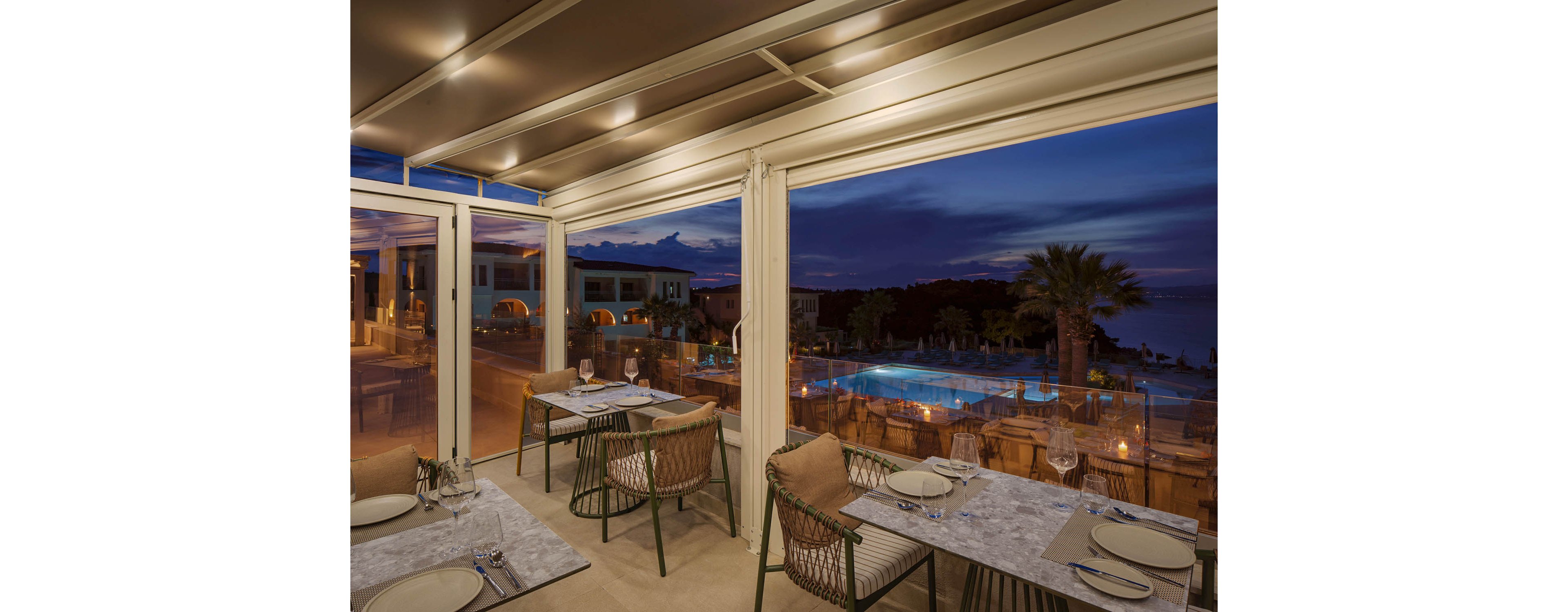 An upscale open-air restaurant overlooks a brightly lit swimming pool at dusk. Tables are neatly set with plates, cutlery, glasses, and placemats, ready for evening diners. Large windows and a covered terrace frame the deep blue sky and distant sea. Warm ambient lighting and modern wicker chairs create a relaxed, sophisticated atmosphere.