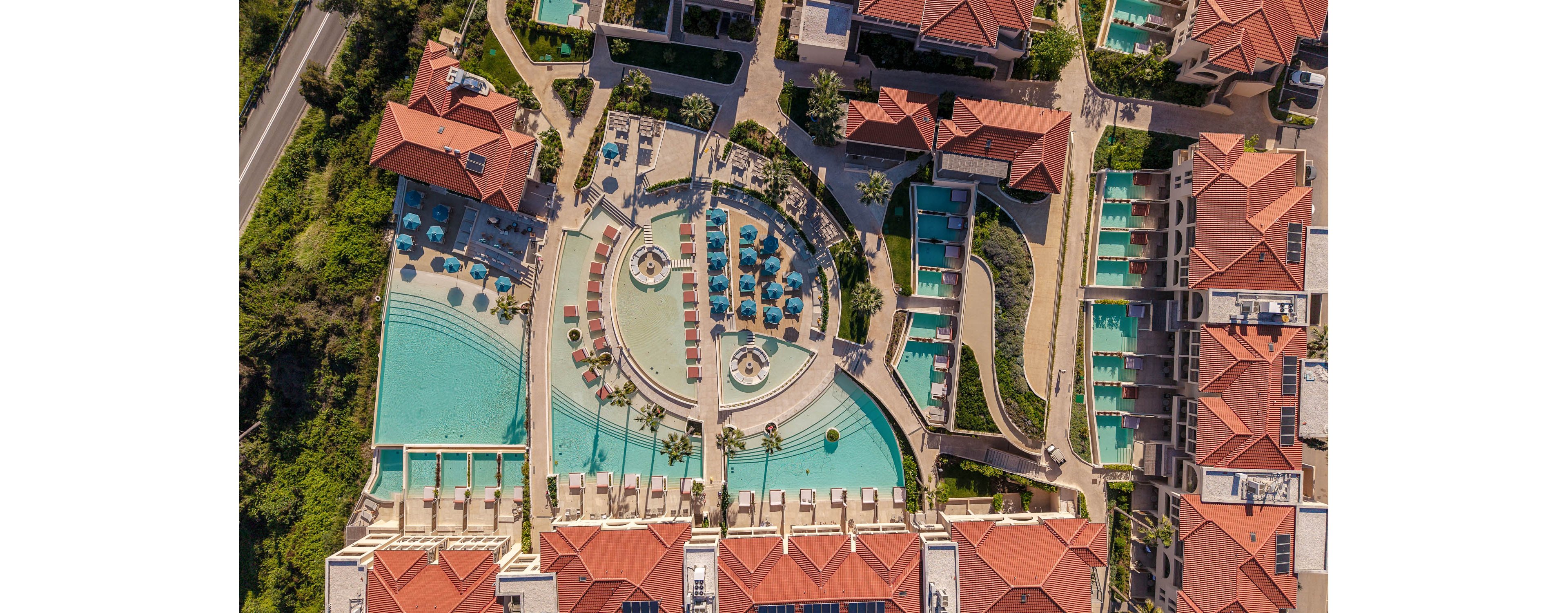 An aerial view captures a large luxury resort complex with multiple swimming pools and terraced sun decks. The central circular pool area is surrounded by lounge chairs and umbrellas, with additional rectangular pools extending along the buildings. Red-tiled roofs and neatly arranged pathways frame the turquoise water, creating a geometric pattern. The scene suggests a sunny, relaxing vacation atmosphere in a coastal or hillside setting.