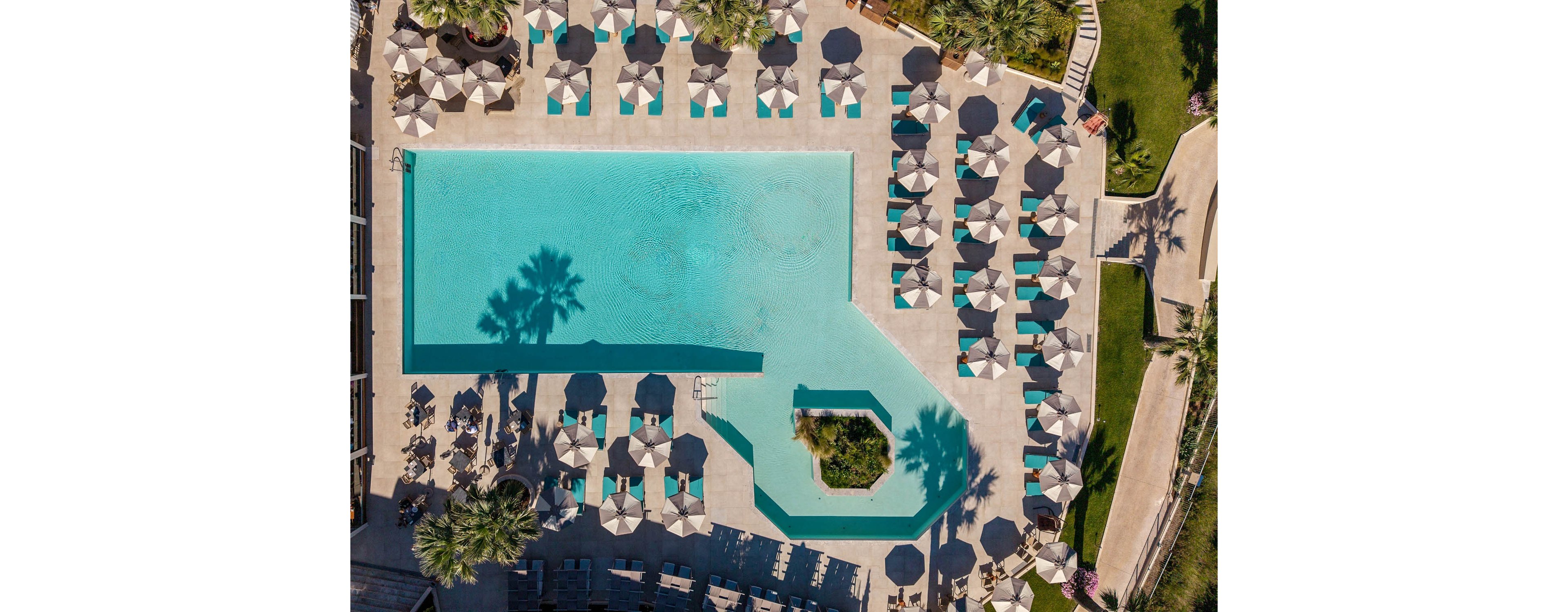 A large resort swimming pool is photographed from directly above, revealing its geometric shape and clear blue water. Numerous sun loungers with matching umbrellas are neatly arranged around the pool on a light-colored terrace. Palm trees cast distinct shadows across the deck, adding visual interest and contrast. The scene suggests a sunny, relaxing vacation atmosphere in a well-maintained outdoor leisure area.