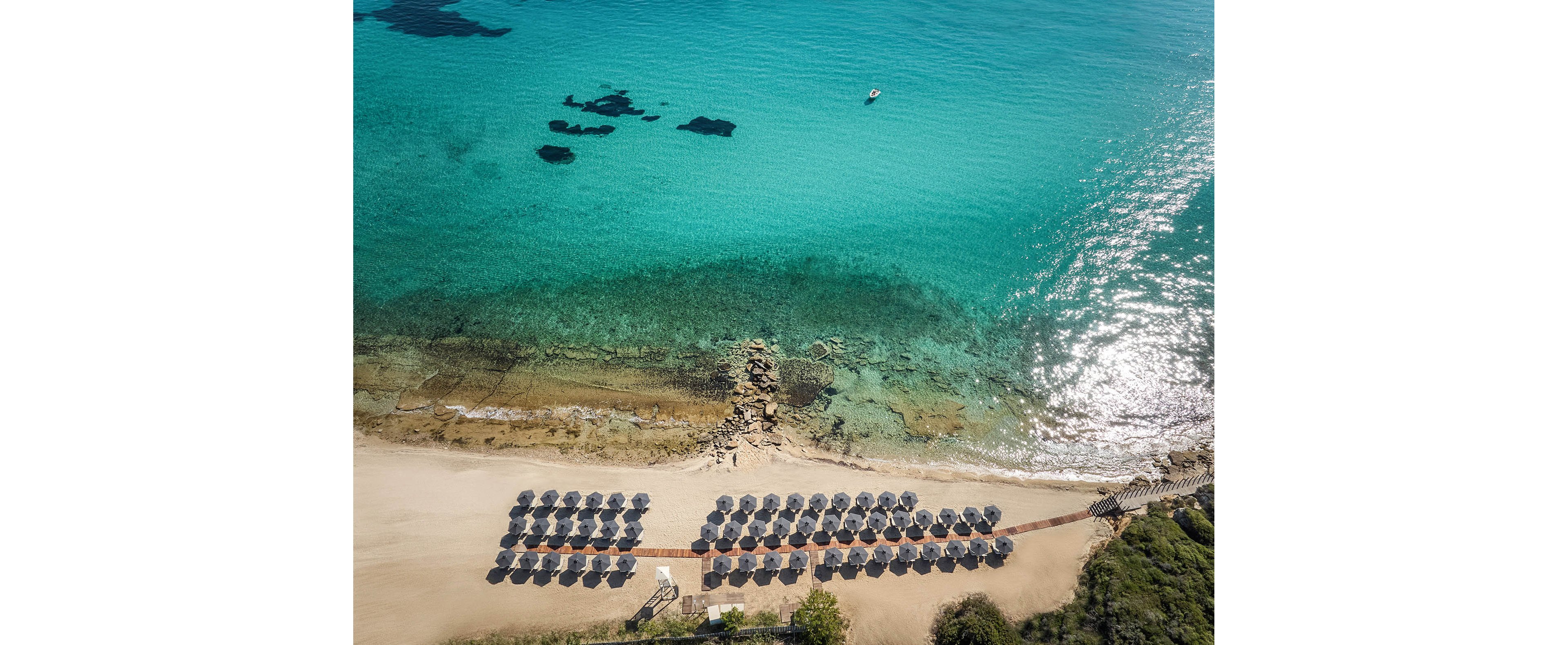 An aerial view shows a neatly arranged row of beach umbrellas on a sandy shoreline. The umbrellas are aligned in multiple rows along a wooden walkway leading toward the sea. Clear turquoise water with visible rocks and shallow areas stretches out from the shore. A small boat floats in the distance, emphasizing the calm and serene coastal setting.