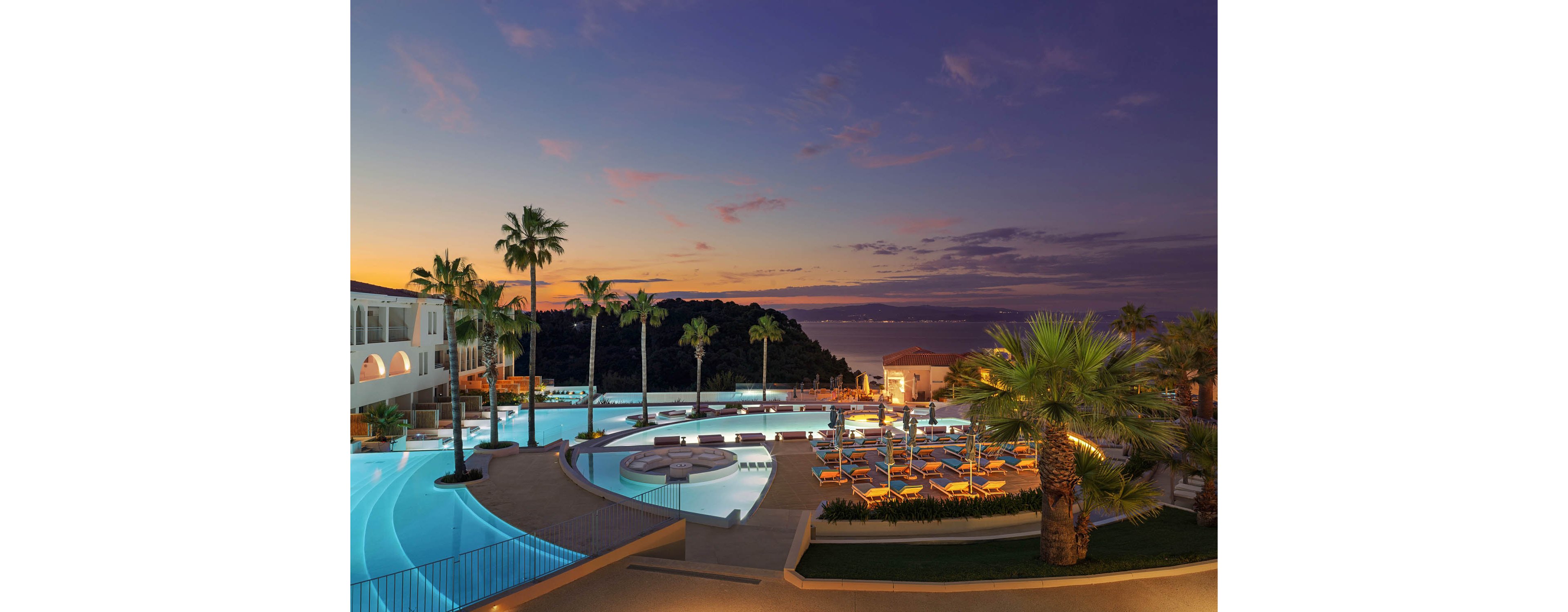 A luxurious seaside resort features a large infinity pool complex overlooking the ocean at dusk. Curved pools, sun loungers, and palm trees create a relaxing vacation atmosphere. Warm lights illuminate the terraces and walkways as the sky glows with soft pink and purple clouds. The distant coastline and calm water enhance the tranquil, tropical setting.
