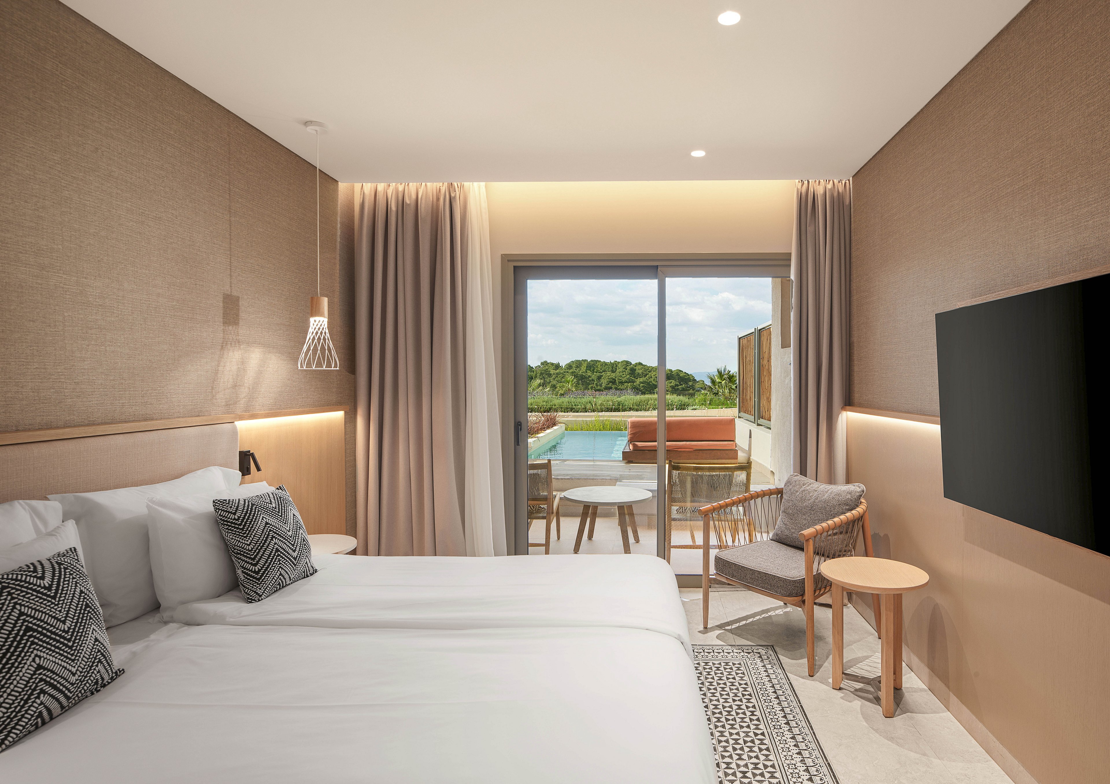A contemporary hotel room features a neatly made double bed with white linens and patterned cushions. Neutral-toned walls, soft curtains, and warm ambient lighting create a calm and inviting atmosphere. A sliding glass door opens to a private terrace with chairs, a small table, and a pool overlooking greenery. A flat-screen television is mounted on the wall opposite the bed, completing the modern interior.