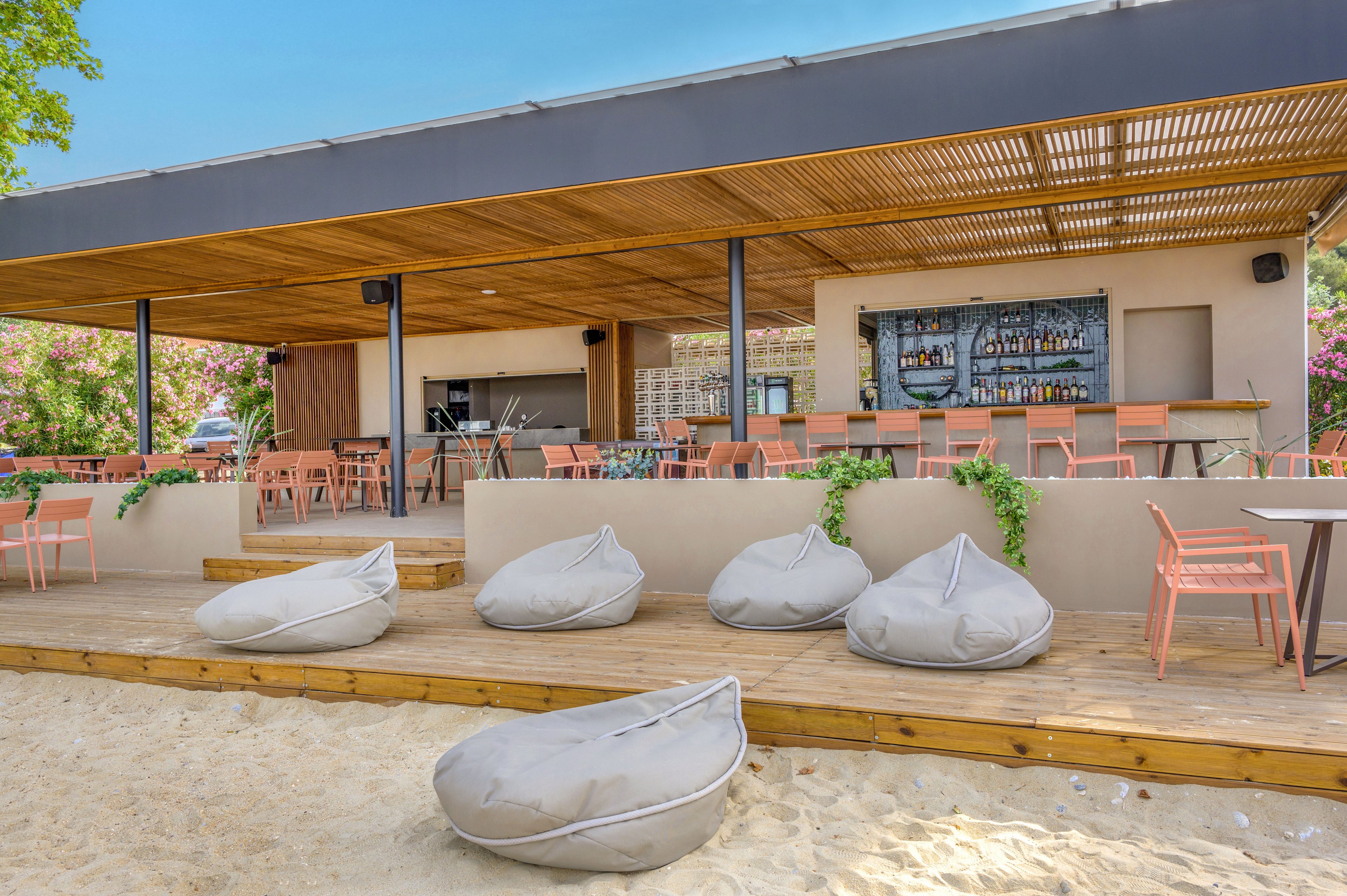 An open-air bar terrace overlooks a sandy beach with several gray beanbag loungers in the foreground. The wooden deck features pastel-colored chairs and tables arranged in front of a long bar counter stocked with bottles and glassware. A slatted wooden roof provides shade, while greenery and flowering bushes add color around the structure. The scene suggests a relaxed coastal setting ideal for casual drinks and socializing.