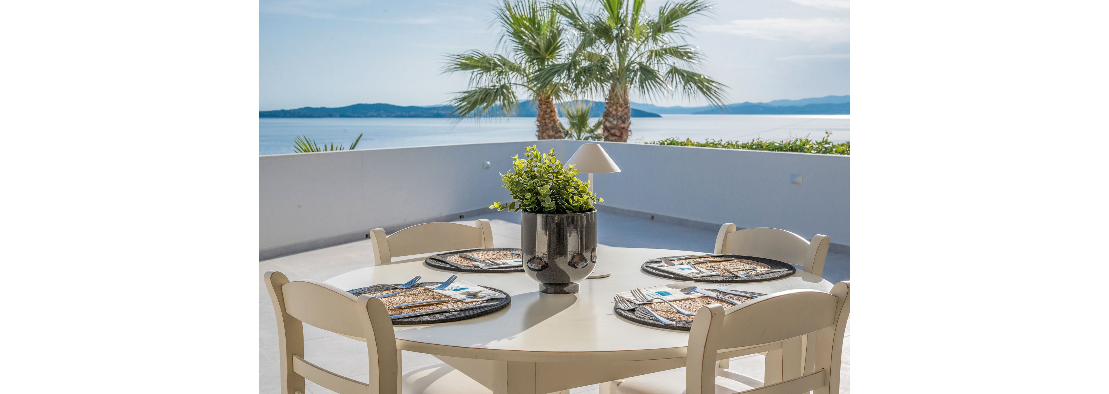 A round outdoor dining table is set on a bright terrace overlooking a calm sea. Four chairs surround the table, which is arranged with placemats, cutlery, and plates ready for a meal. A shiny black planter with green foliage sits at the center, accompanied by a small modern lamp. In the background, tall palm trees frame the coastal landscape under a clear blue sky.
