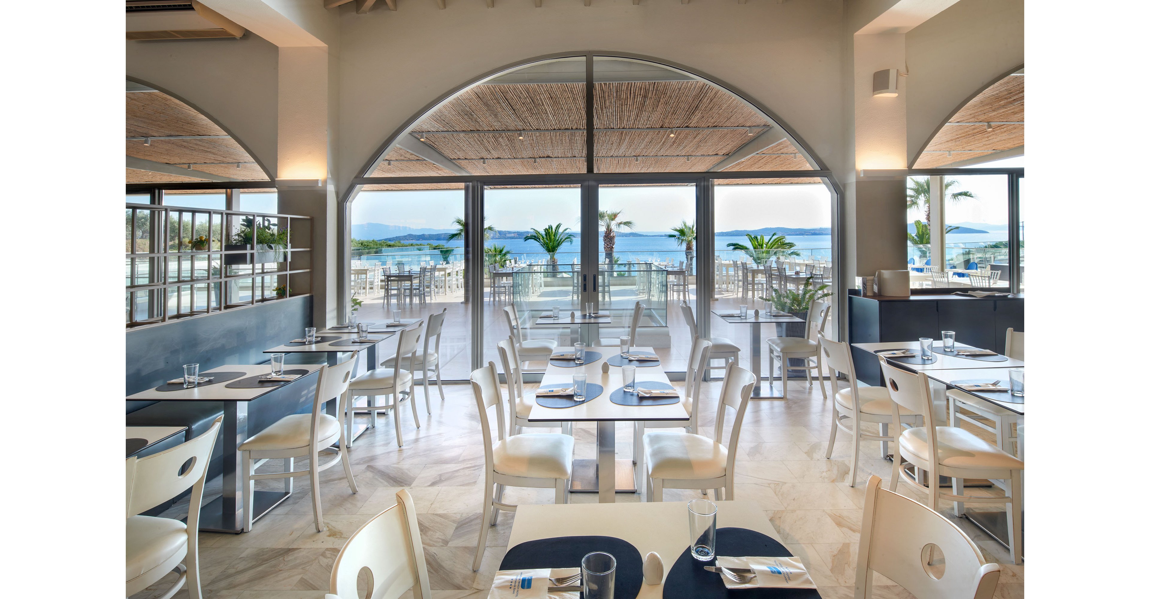Contemporary restaurant interior features neatly arranged tables and white chairs facing large windows. The space opens onto an outdoor terrace with additional seating and views of the ocean and palm trees. Natural light floods the room, highlighting the neutral tones, blue accents, and clean, minimalist decor. The setting feels calm and inviting, ideal for relaxed coastal dining.