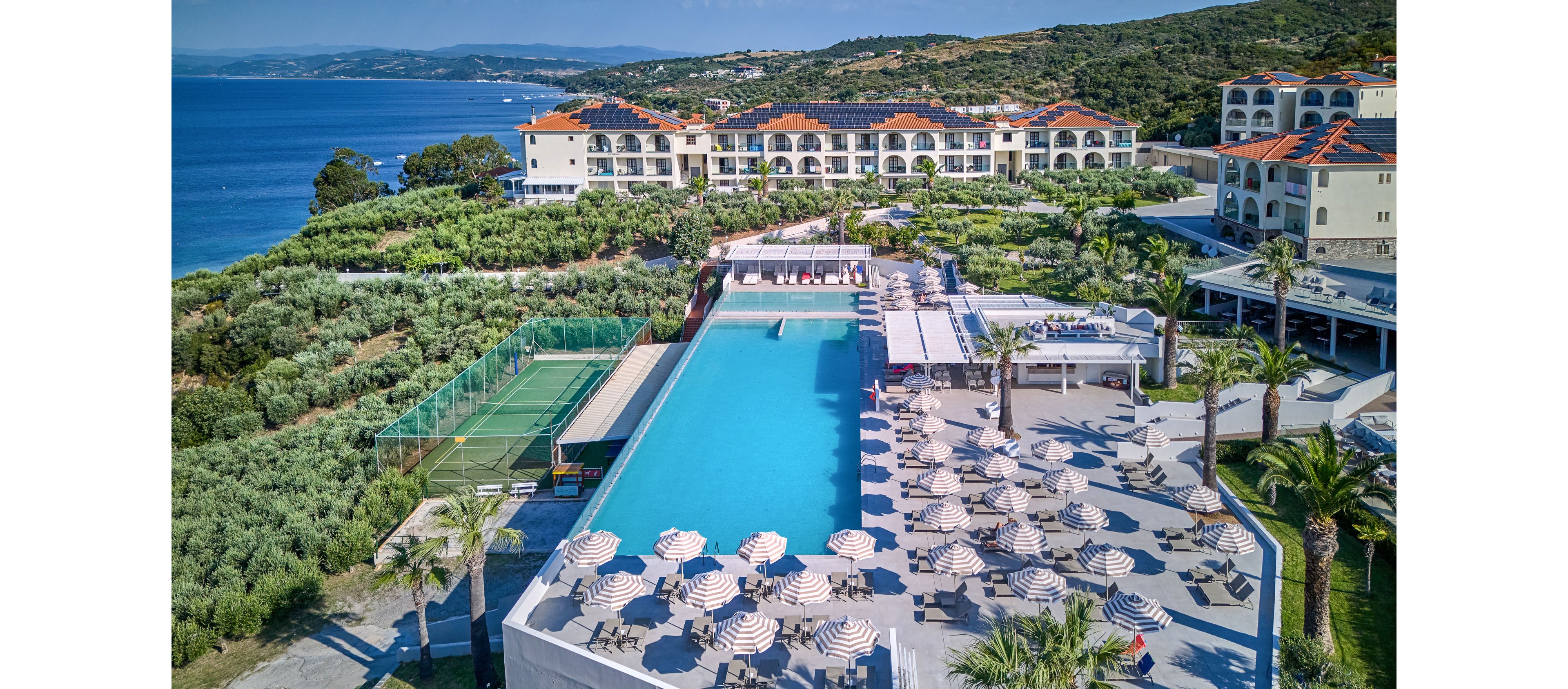 A large seaside resort features an expansive infinity pool overlooking the ocean. Numerous sun loungers and striped umbrellas line the poolside terrace, creating a relaxed vacation atmosphere. The property includes landscaped gardens, palm trees, and a nearby sports court surrounded by greenery. In the background, multi-story hotel buildings with red-tiled roofs face the coastline and distant hills under a clear blue sky.