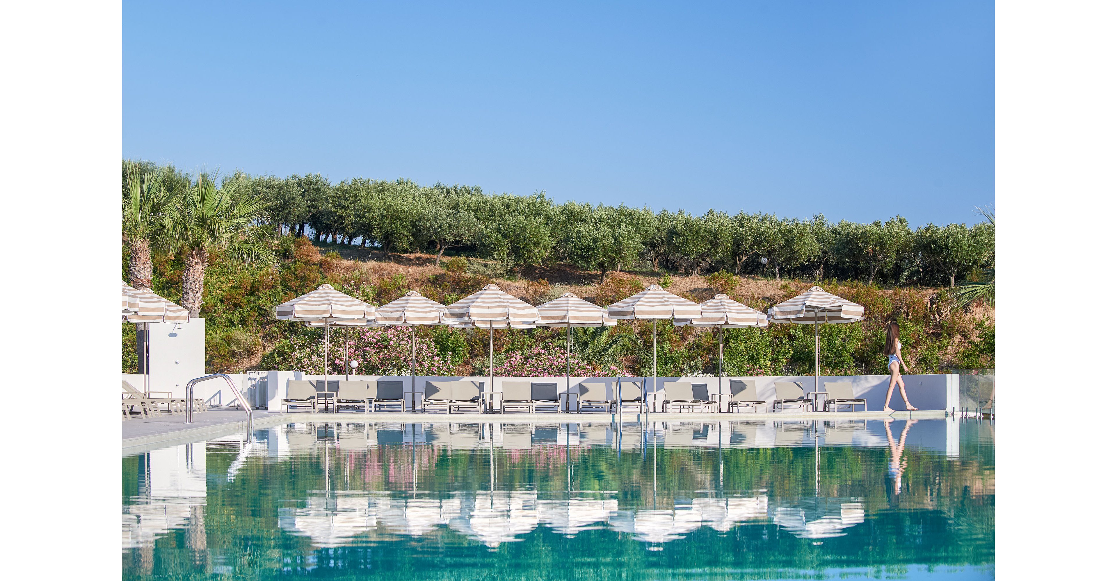 A large outdoor swimming pool reflects rows of striped sun umbrellas and loungers along its edge. A person walks beside the pool, adding a sense of scale and quiet activity to the serene resort setting. Lush greenery, palm trees, and flowering shrubs rise behind the pool area under a clear blue sky. The overall scene conveys a calm, sunny vacation atmosphere with clean, modern design.