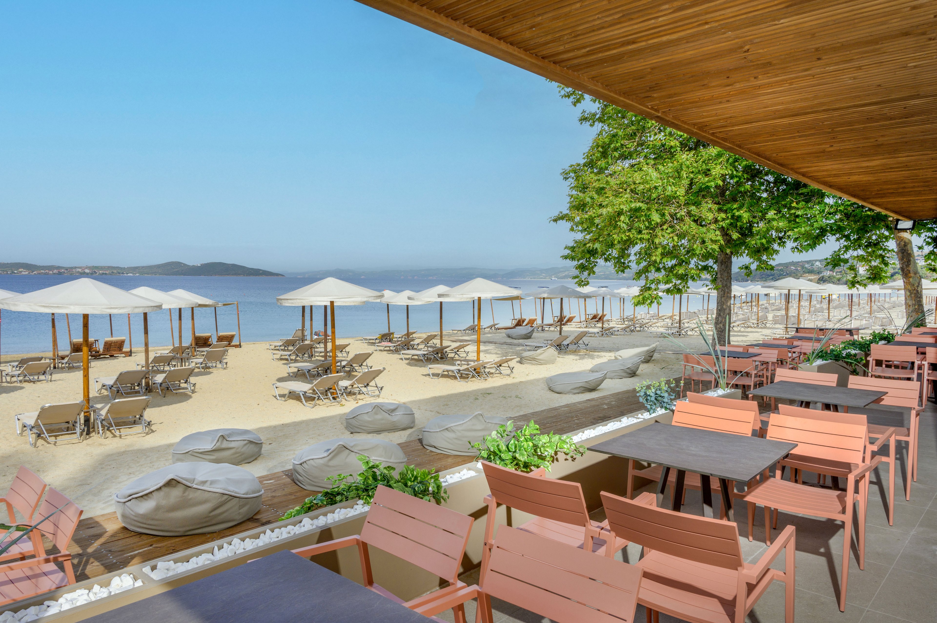 An open-air café terrace with wooden tables and chairs faces a sandy beach lined with sun loungers and white umbrellas. Bean bag seats and low plants separate the dining area from the shore, creating a relaxed coastal atmosphere. Calm blue water stretches into the distance under a clear sky, with gentle hills visible on the horizon. The scene suggests a tranquil seaside resort ideal for outdoor dining and beach relaxation.