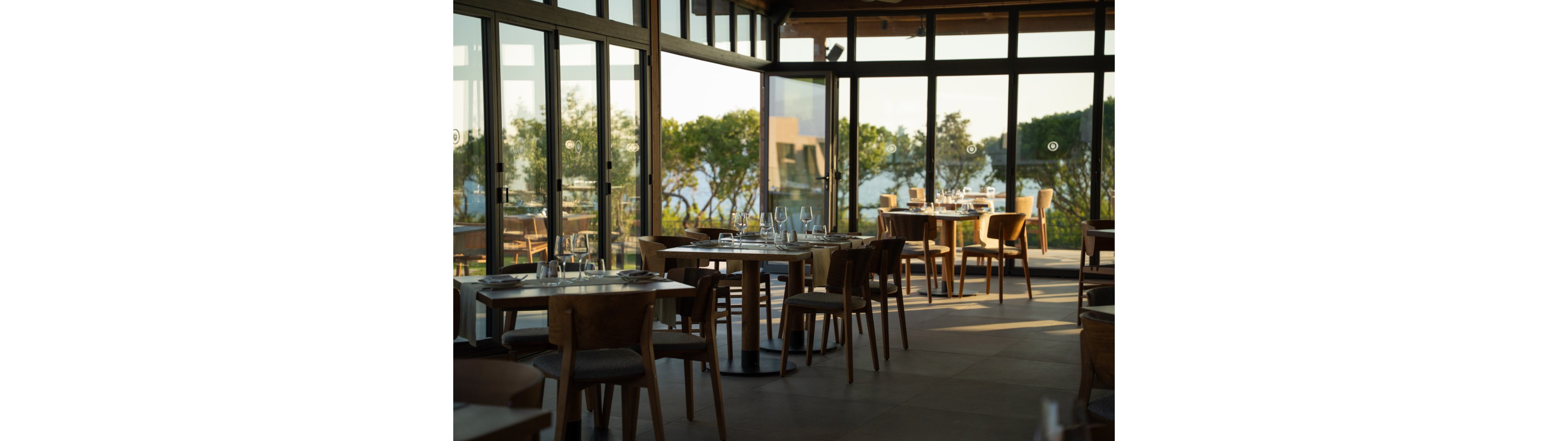 A modern restaurant dining area is shown with neatly arranged wooden tables and chairs. Each table is set with glassware and cutlery, ready for guests. Large floor-to-ceiling windows surround the space, offering views of trees and water outside. Warm natural light fills the interior, creating a calm and inviting atmosphere.