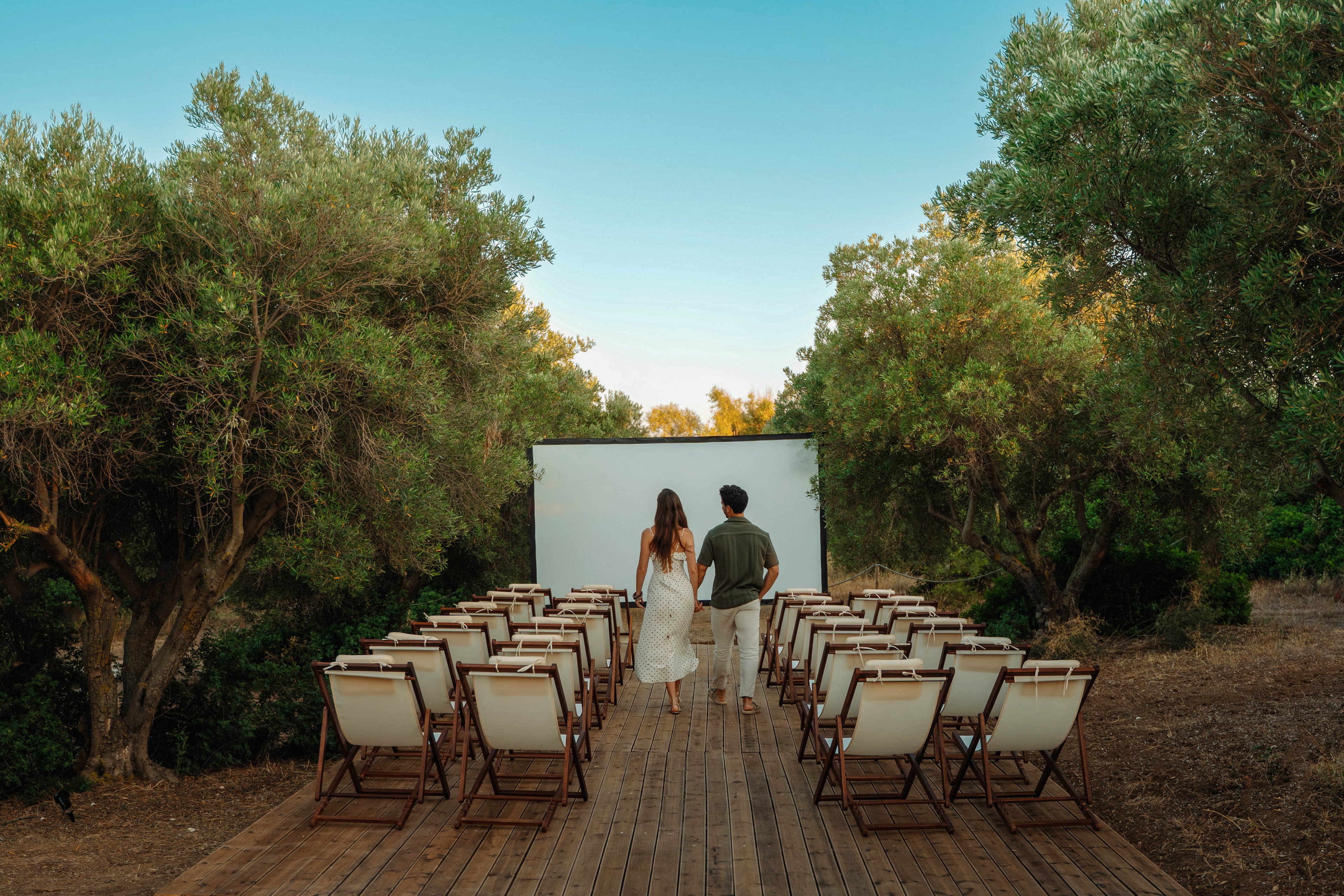 A couple walks hand in hand toward a large white screen set up outdoors. Rows of empty lounge-style chairs line a wooden deck, creating an intimate open-air cinema or event space. Tall green trees surround the area under a clear blue sky, giving the scene a secluded, natural feel. The overall atmosphere is calm, romantic, and anticipatory.