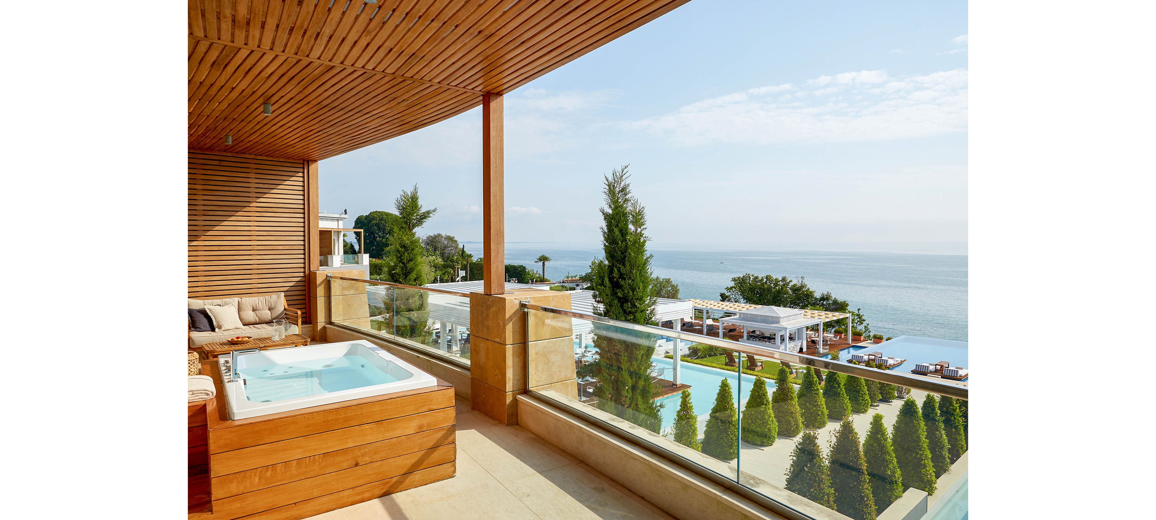 A modern terrace features a built-in hot tub framed with warm wooden paneling. Glass railings open the space to a panoramic view of the sea and distant horizon. Below, manicured trees line a resort pool area with white cabanas and sun loungers. The scene is bright and tranquil, evoking a luxurious coastal vacation atmosphere.