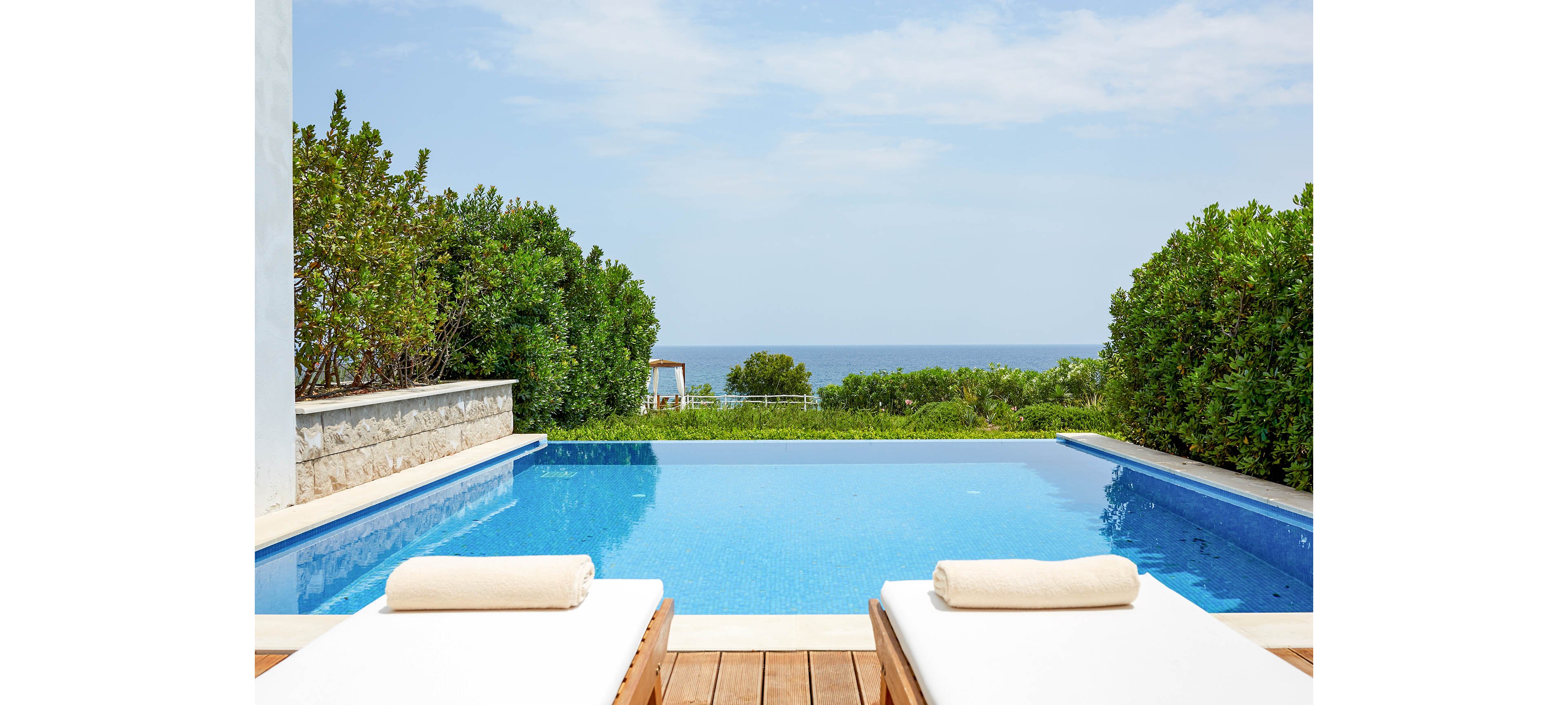 A serene infinity pool overlooks a distant ocean horizon on a clear day. Two white cushioned loungers with folded towels sit on a wooden deck in the foreground. Lush green shrubs and a stone wall frame the pool, creating a private, resort-like atmosphere. The scene feels calm and inviting, ideal for relaxation and summer vacations.