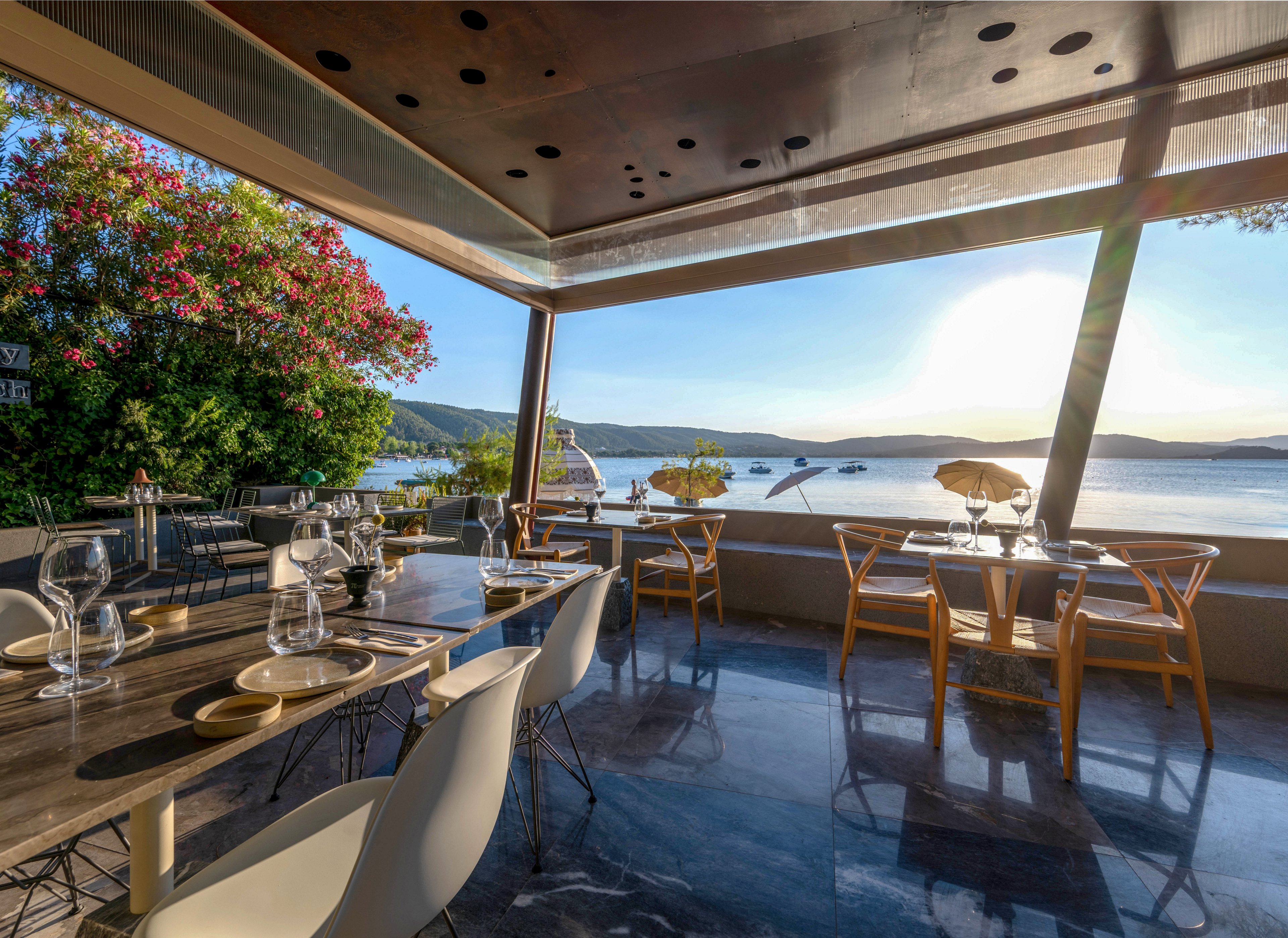 Elegant outdoor restaurant seating overlooks a calm waterfront at sunset. Glass-topped tables are set with wine glasses, plates, and cutlery, ready for guests. Wooden and modern white chairs face the scenic bay with boats and distant hills. Warm sunlight floods the terrace, reflecting on the polished floor and creating a relaxed, upscale atmosphere.