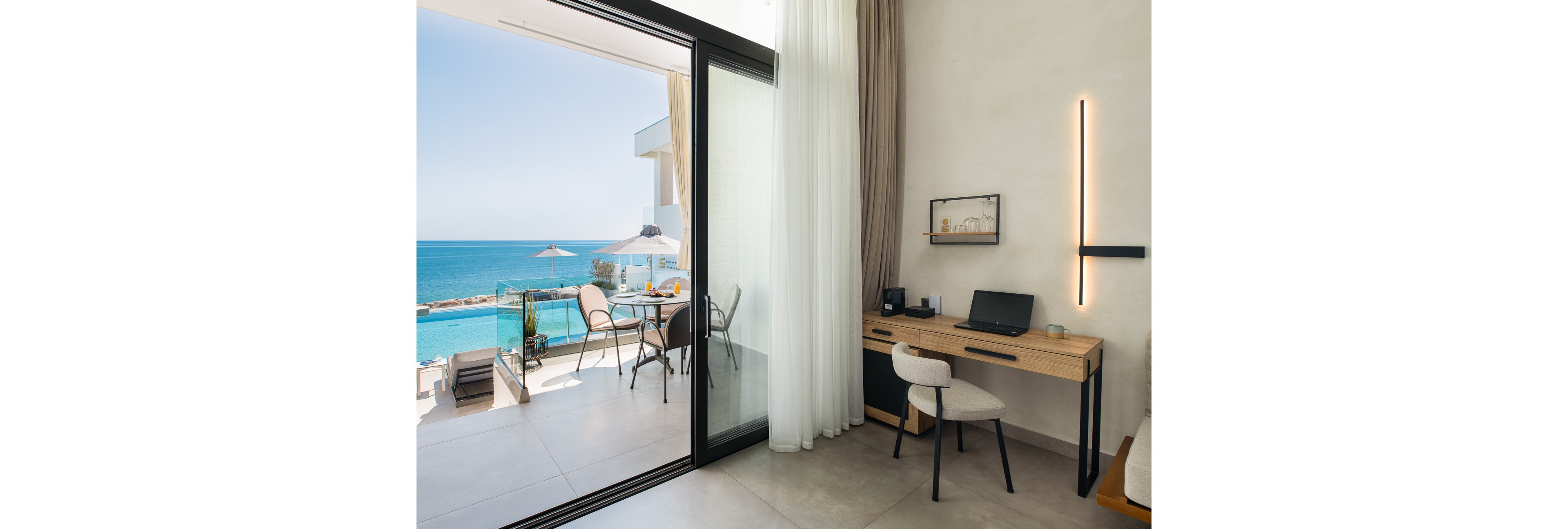 A contemporary hotel room features a minimalist wooden desk and chair set up as a workspace. Large sliding glass doors open onto a balcony with a table, chairs, and sun umbrella overlooking a bright blue sea. Sheer and opaque curtains frame the doorway, while a slim vertical wall light adds a modern touch. The scene feels calm and inviting, ideal for remote work or relaxation by the water.