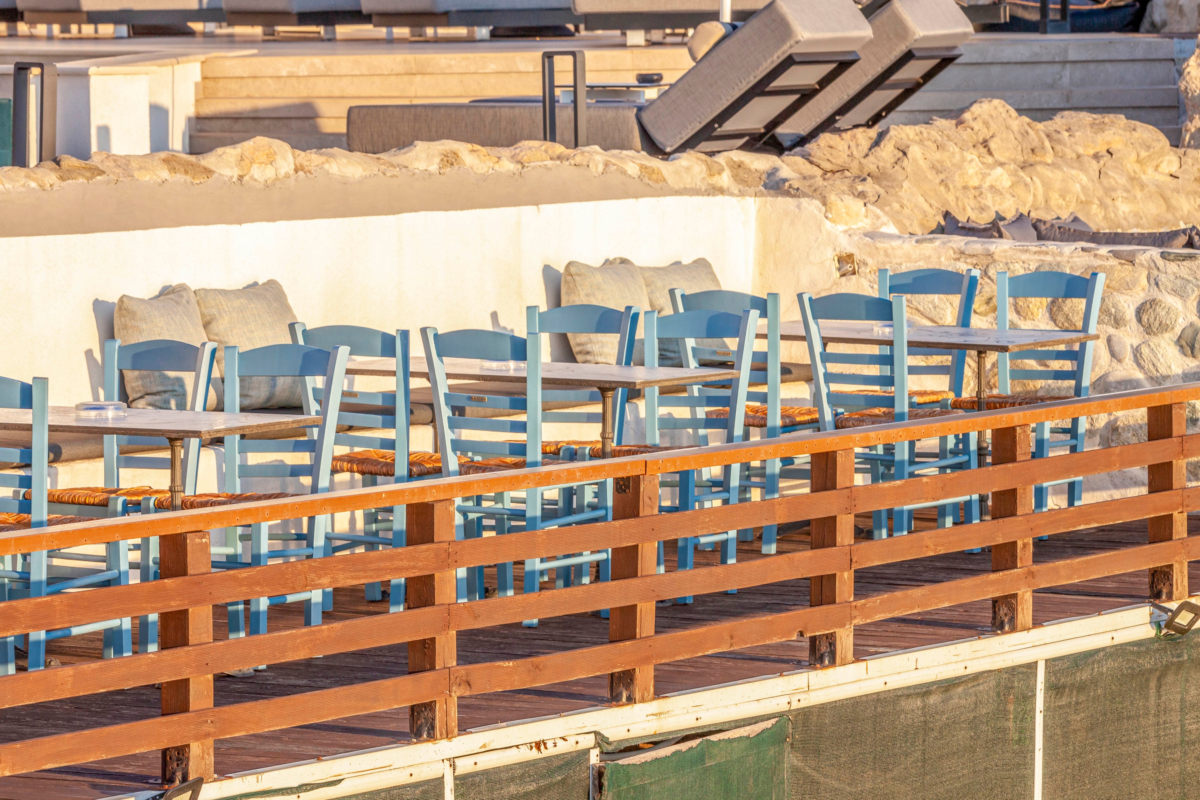 The image shows an outdoor seating area featuring several blue wooden chairs and tables arranged along a terrace. Cushions are placed on the chairs, and the setting is bathed in warm sunlight, suggesting late afternoon or early evening. The terrace is bordered by a wooden railing and stone wall, creating a relaxed and inviting atmosphere. No visible text or numeric values are present.