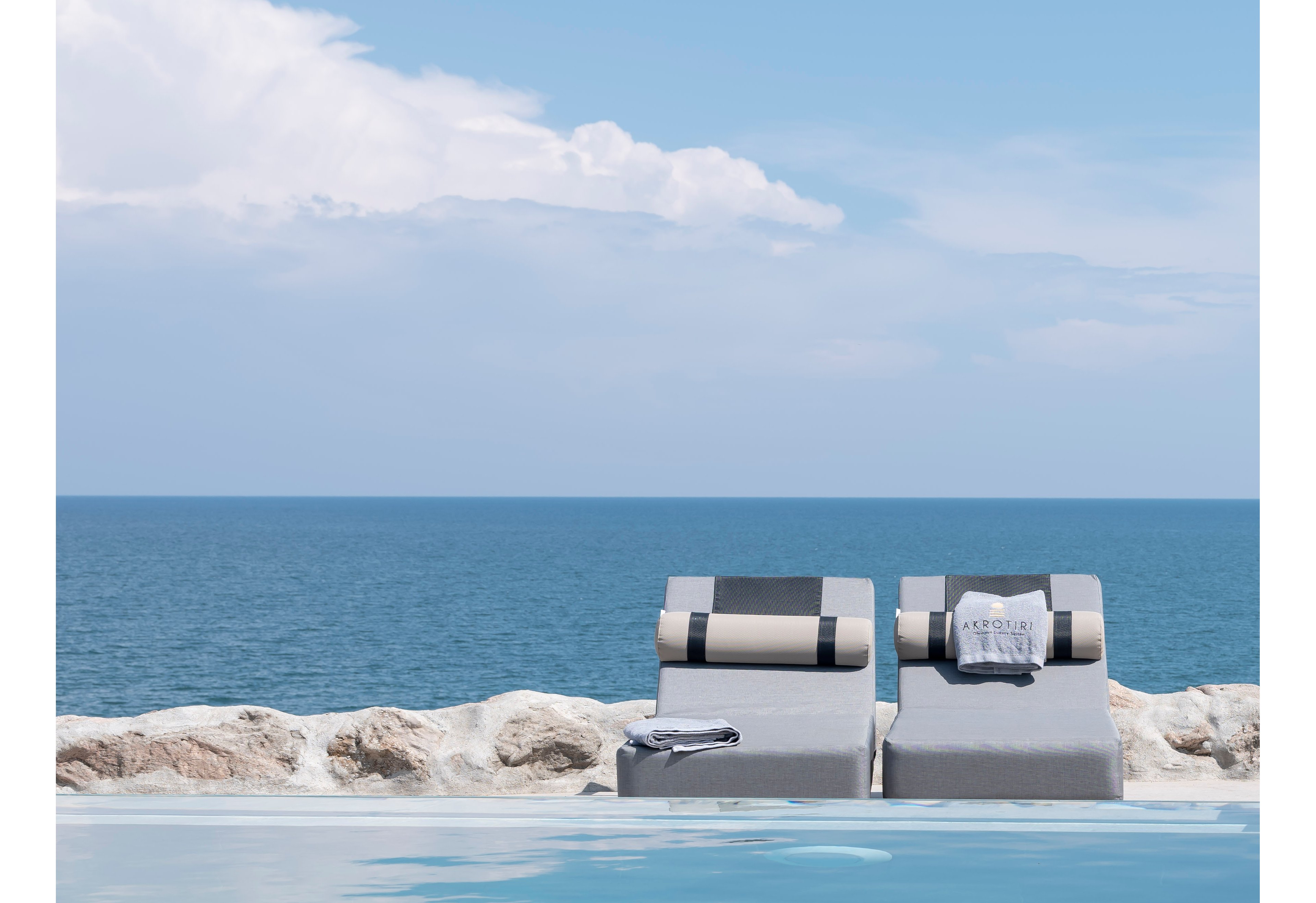 Two modern lounge chairs are positioned beside a serene infinity pool, facing a vast blue ocean under a partly cloudy sky. One lounger features a rolled towel with visible text reading 'ACQUA', adding a touch of sophistication. The setting evokes relaxation and leisure, with neutral tones and minimalist style dominating the scene. No people are present, emphasizing tranquility and exclusivity.