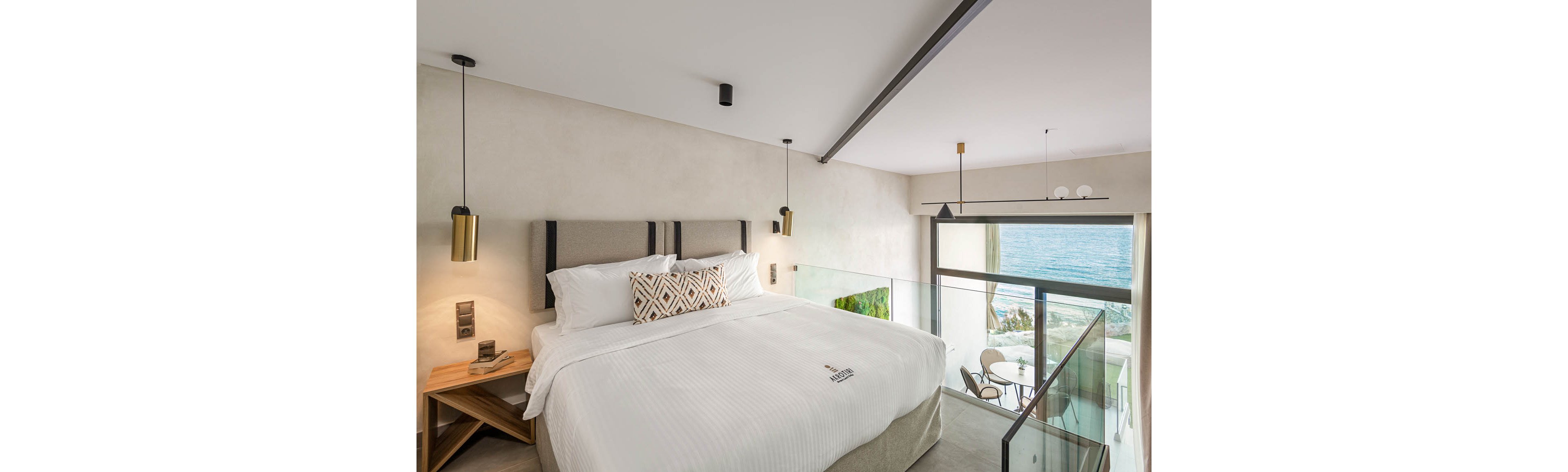A contemporary hotel room features a large bed with crisp white linens and a geometric accent pillow. The space is illuminated by stylish pendant lights and natural sunlight streaming through floor-to-ceiling windows. Outside, a private balcony overlooks the ocean, furnished with a small table and chairs. The visible text on the bed reads 'Minos Beach Art Hotel.'