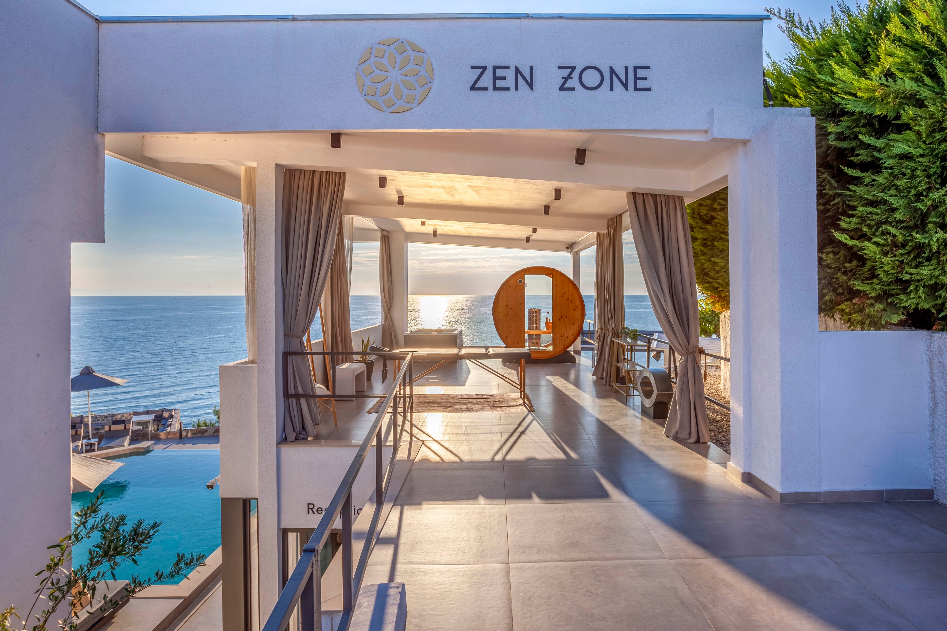 A contemporary open-air lounge labeled 'ZEN ZONE' overlooks a tranquil ocean view. The setting features elegant white architecture, flowing curtains, and a circular wooden structure at the far end. The scene is bathed in warm sunlight, evoking a serene and relaxing atmosphere, with a pool and sun loungers visible below.