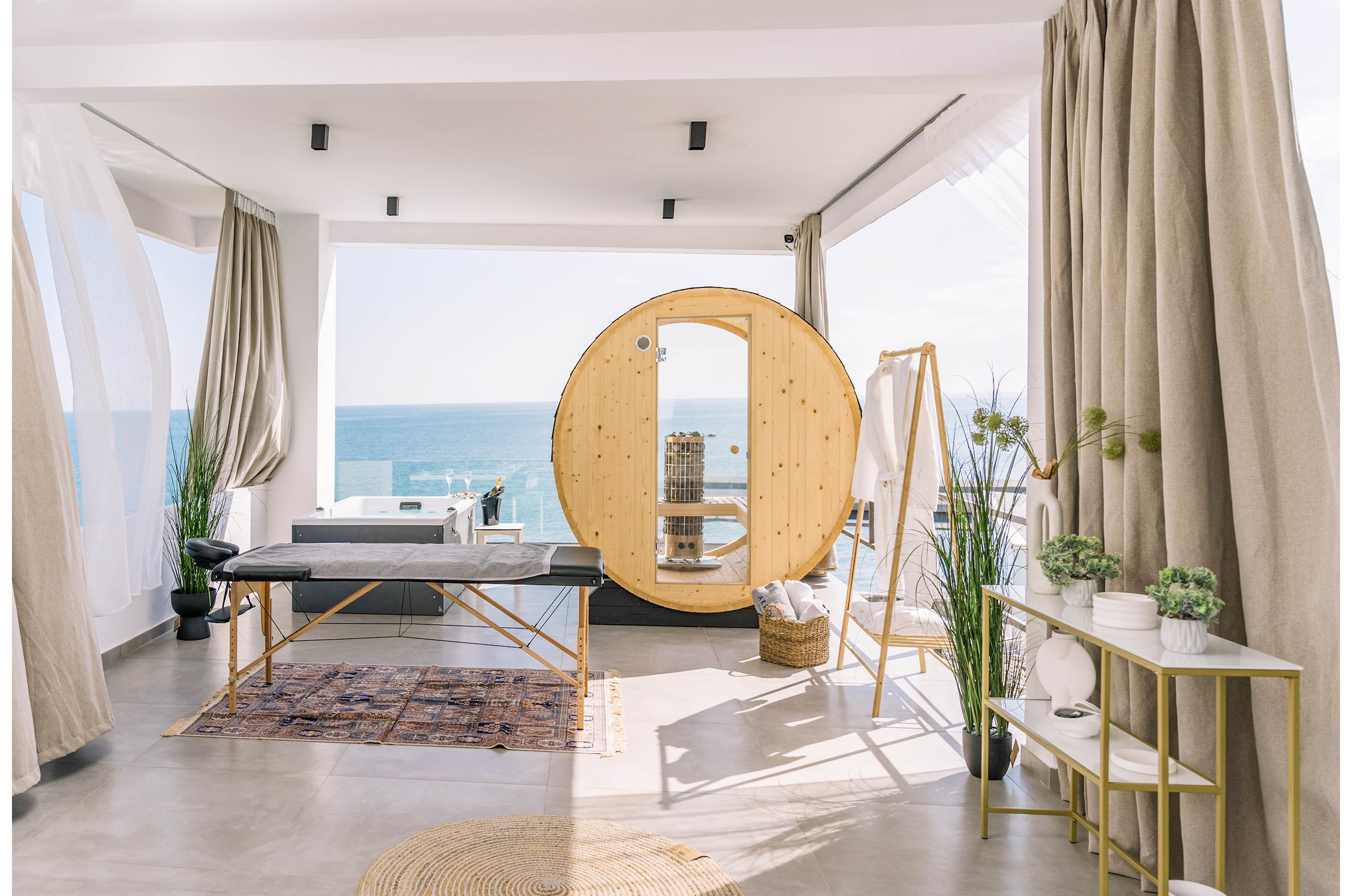 A contemporary spa room features a circular wooden sauna and a massage table, set against a panoramic ocean backdrop. The space is decorated with neutral tones, natural light, and minimalist furnishings, including plants and woven baskets. Sheer curtains and soft textiles create a tranquil, airy atmosphere, ideal for relaxation and wellness. No visible numeric values or identifiable brands are present.