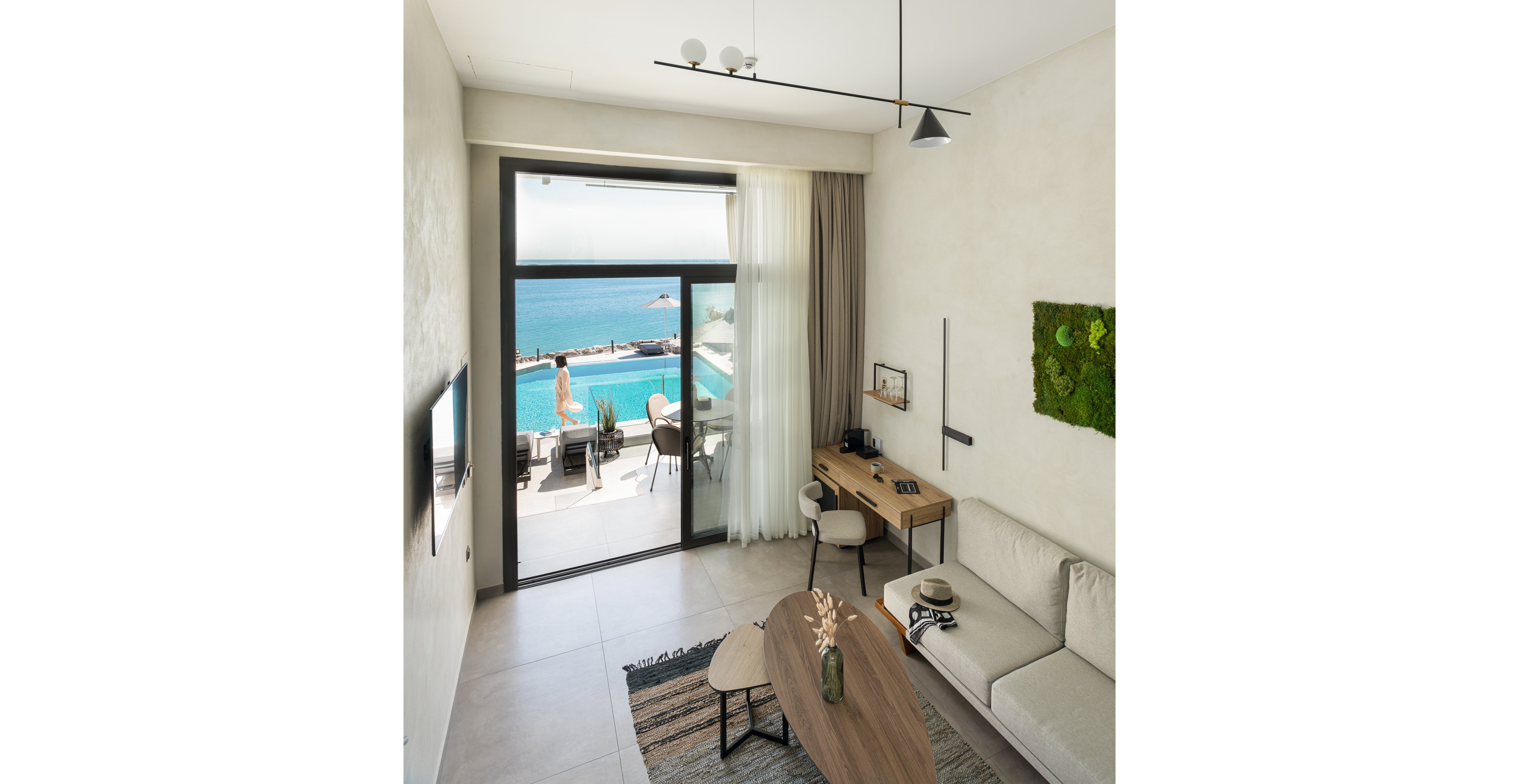 A contemporary hotel suite features a cozy living area with neutral-toned furniture and a workspace. Large glass doors open to a private terrace overlooking a turquoise infinity pool and the ocean, where a person is seen relaxing outdoors. The room is decorated with minimalist accents, natural light, and a green wall art piece, creating a serene and inviting atmosphere.