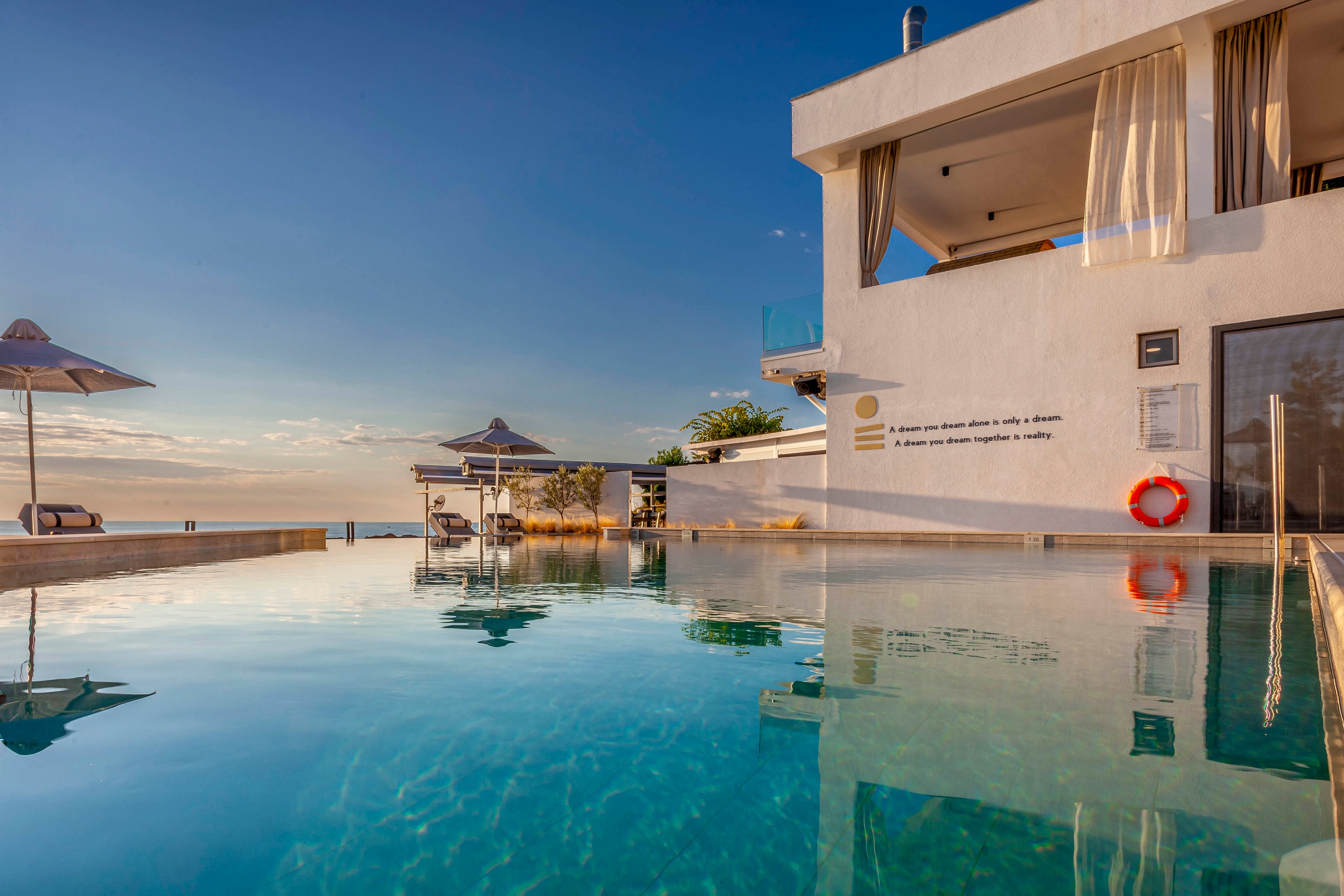 A contemporary villa features a spacious outdoor swimming pool reflecting the clear blue sky. The setting is tranquil, with sun umbrellas and lounge chairs positioned near the water, overlooking the sea. The architecture is minimalist, with white walls and large windows, and a visible safety ring adds a touch of practicality. The mood is serene and inviting, ideal for relaxation and leisure.
