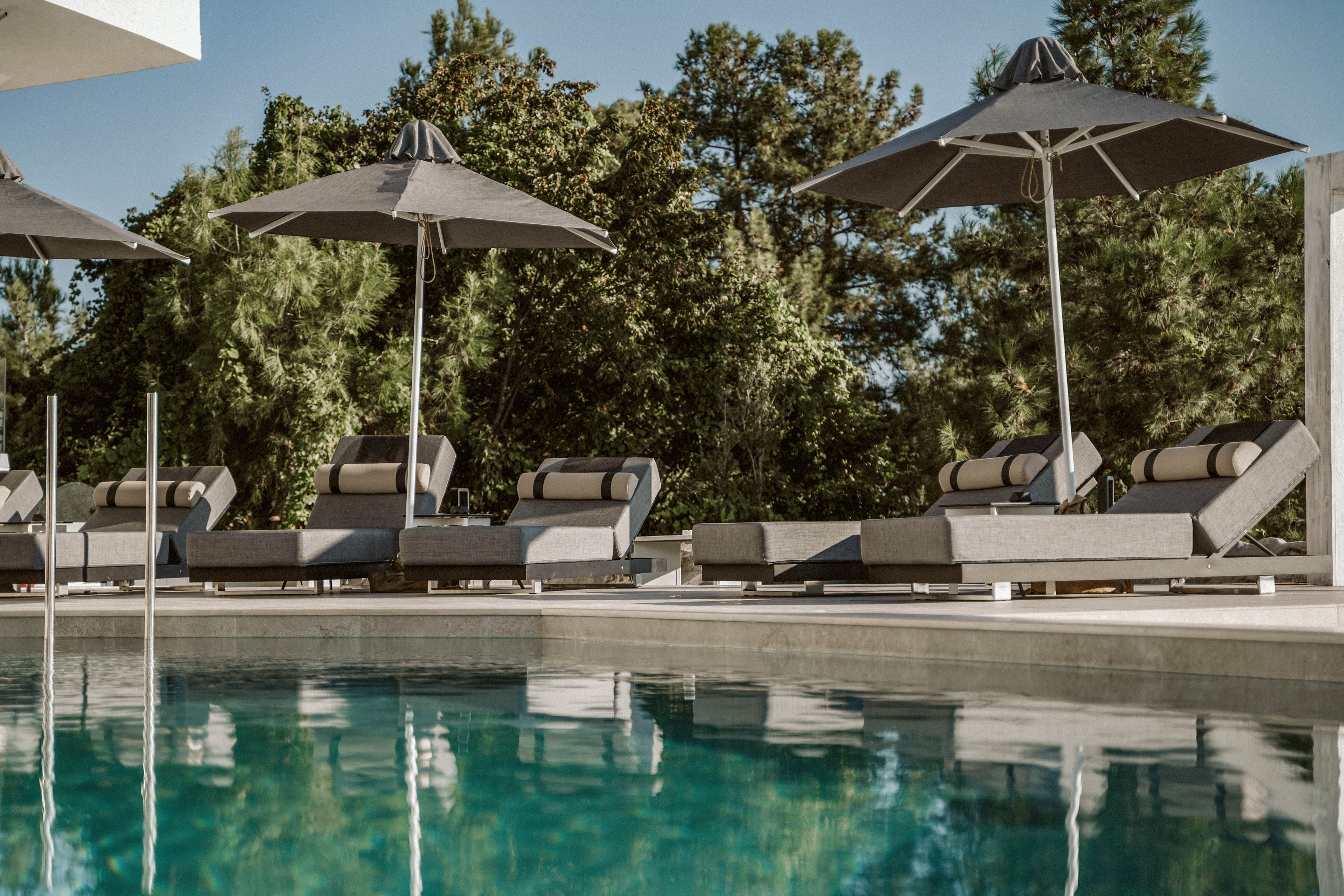 A serene outdoor pool area features several modern lounge chairs arranged in a row, each shaded by a large dark umbrella. The setting is surrounded by lush green trees, creating a tranquil and inviting atmosphere. The water in the pool reflects the clear sky and the poolside furniture, emphasizing a calm and relaxing mood. No people are present, and the color palette is neutral with soft grays and greens.
