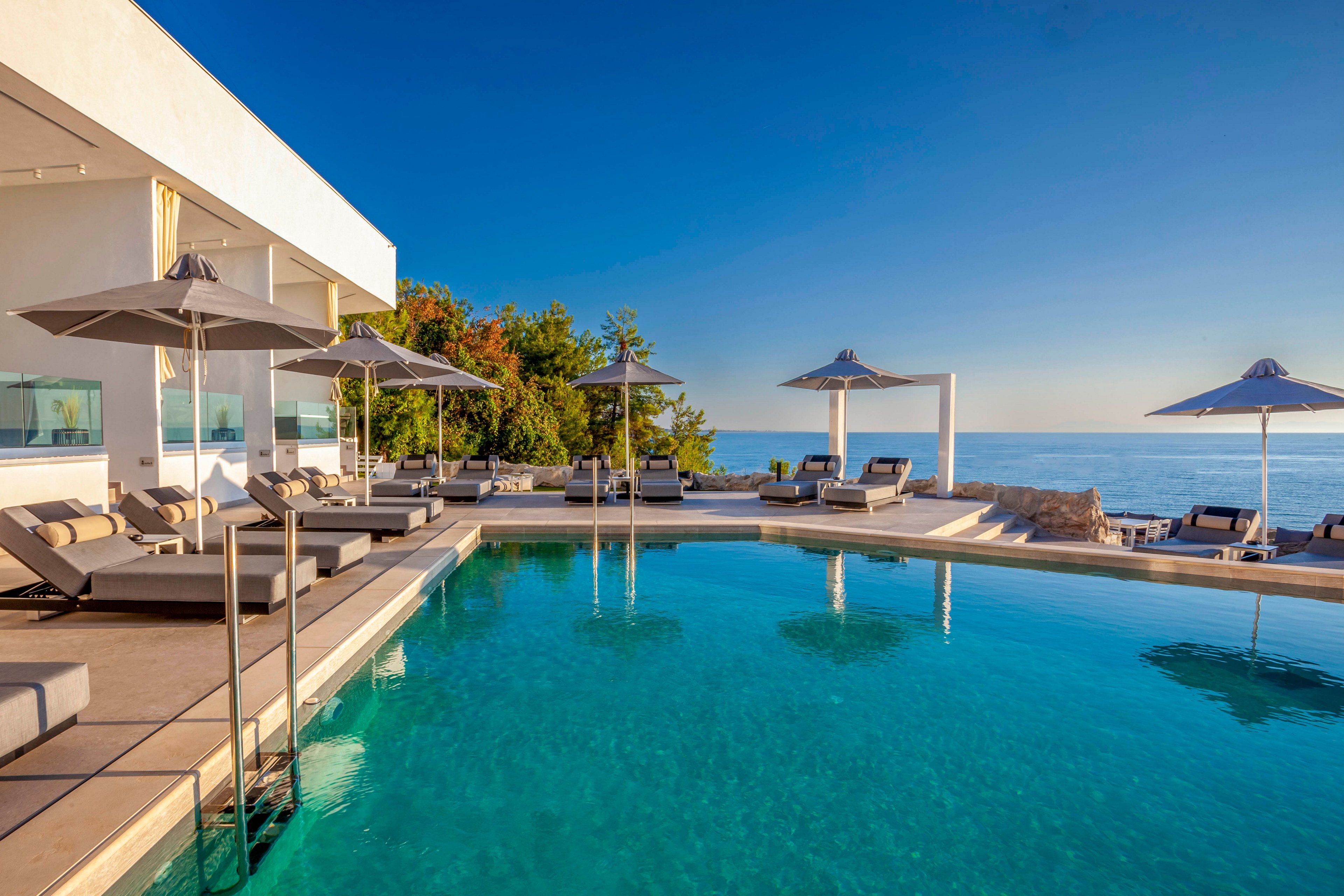 The image features a contemporary outdoor pool area with stylish lounge chairs and umbrellas, set against a backdrop of clear blue skies and calm sea. The setting is tranquil and inviting, ideal for relaxation or sunbathing. The architecture is sleek and minimal, with neutral tones and clean lines enhancing the serene mood. No visible text or numeric values are present in the image.
