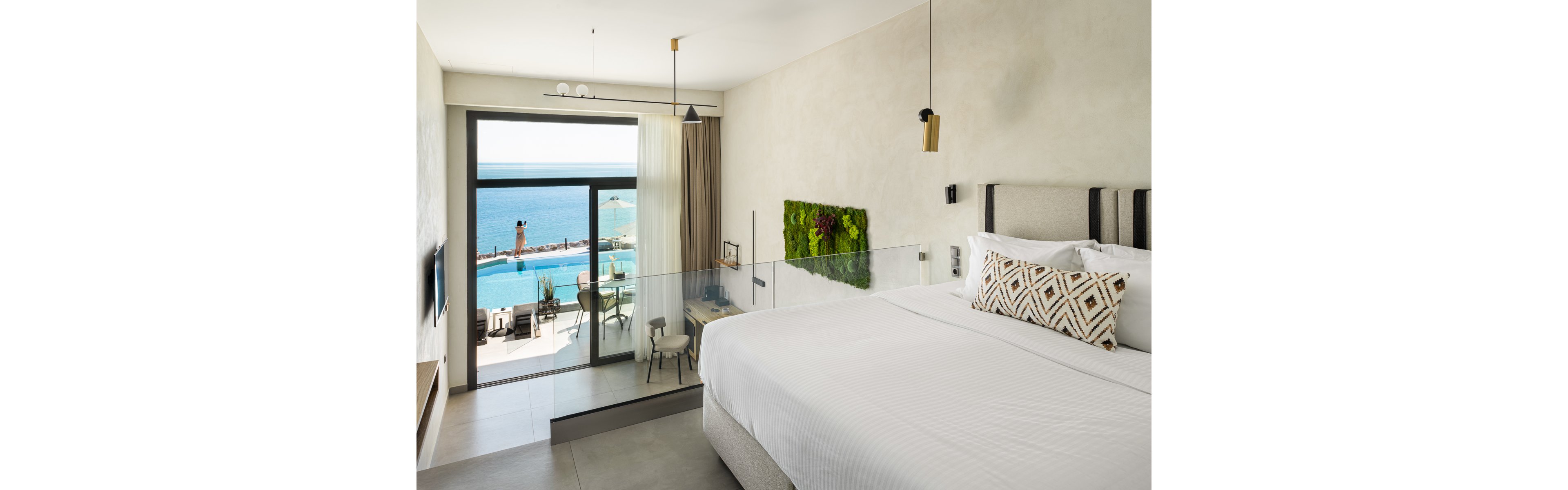 A contemporary hotel room features a large bed with geometric pillows, minimalist decor, and a glass partition. The room opens to a balcony overlooking a pool and the ocean, with a person visible outside. Natural light fills the space, highlighting neutral tones and a green wall accent.