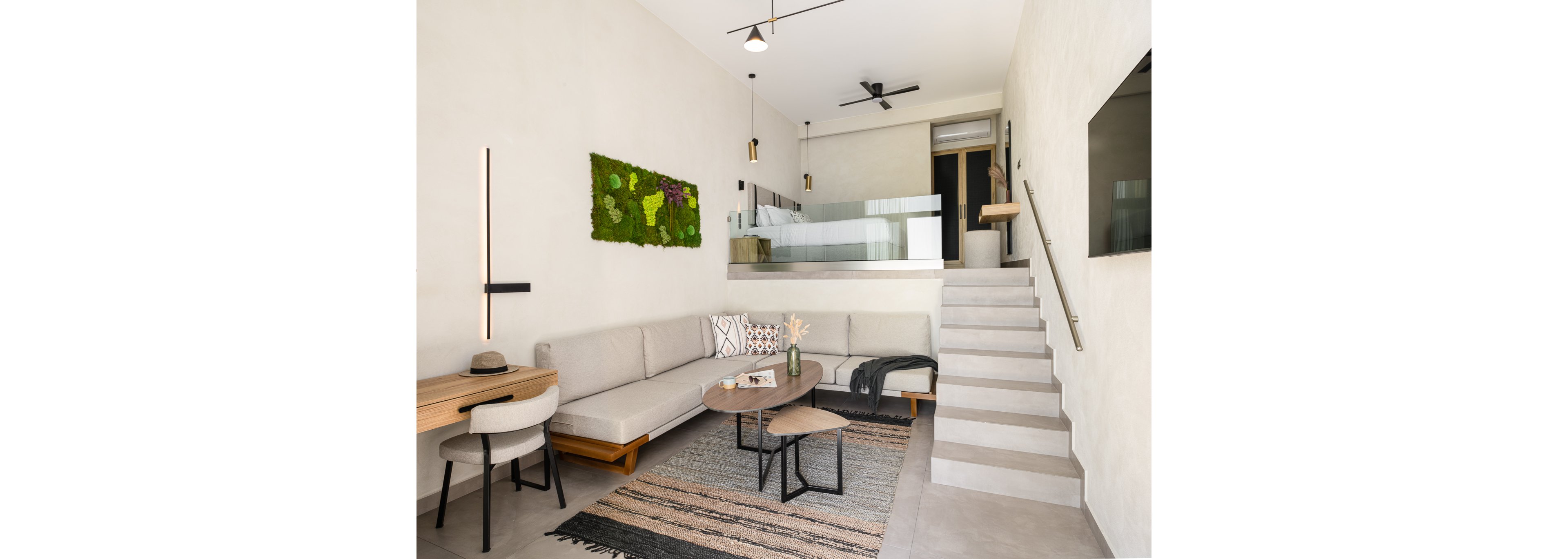 A contemporary living room features a neutral palette with a sectional sofa, wooden coffee tables, and a desk with a chair. The space is accented by a green wall art piece and geometric lighting fixtures. A staircase leads to a loft area with glass railing, and the overall mood is calm and inviting. No visible text or numbers are present in the image.