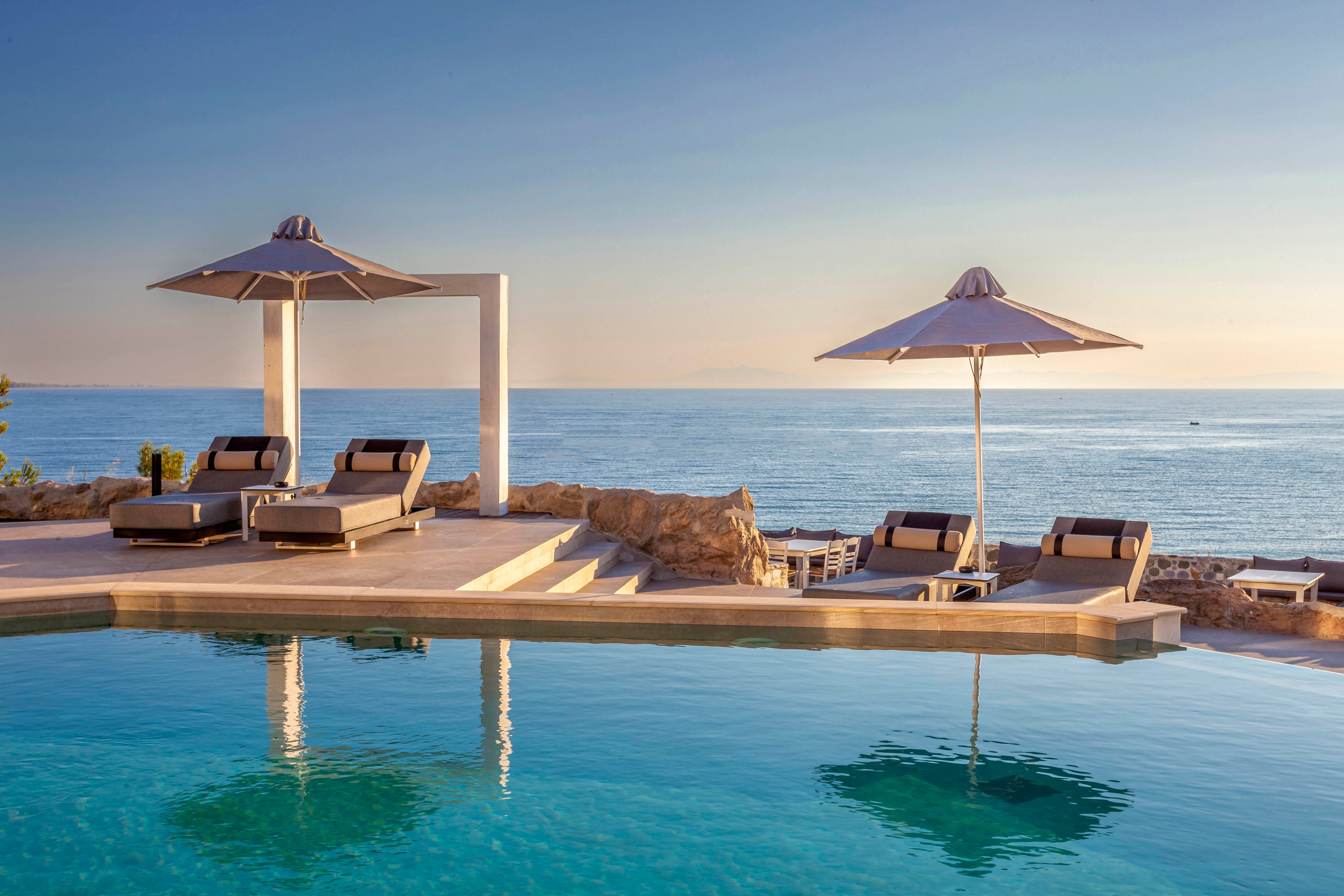 The image features several modern sun loungers and large umbrellas arranged beside an infinity pool, overlooking a calm ocean. The setting is serene and tranquil, with soft sunlight casting gentle reflections on the water. The scene evokes a sense of relaxation and leisure, with neutral tones and a minimalist style. No people are present, emphasizing the peaceful atmosphere.