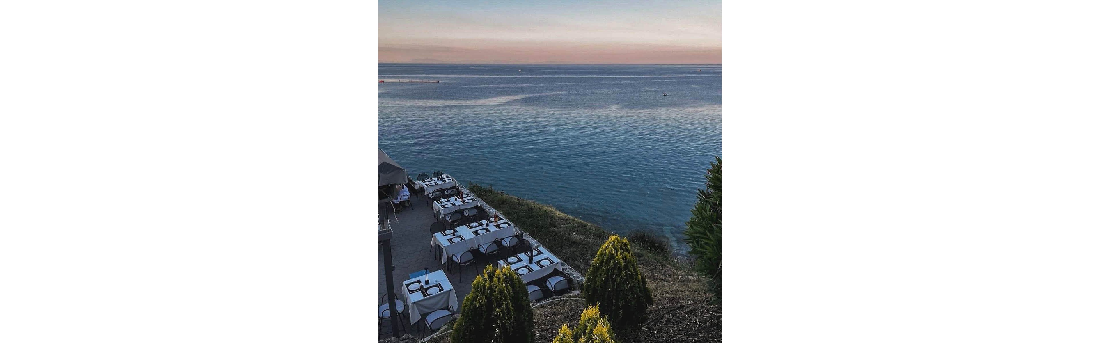 A coastal restaurant terrace features neatly arranged tables with white linens and place settings, overlooking a calm sea at dusk. The scene captures the tranquil ambiance of outdoor dining, with the horizon painted in soft pastel hues. Green shrubs frame the foreground, enhancing the natural beauty and inviting atmosphere. No visible text or numeric values are present in the image.