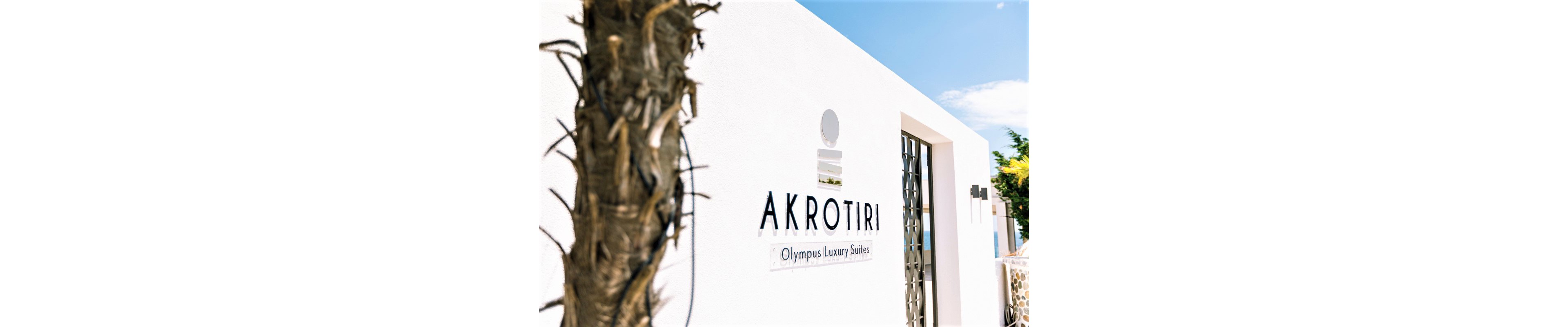 The main subject is the entrance of Akrotiri Olympus Luxury Suites, featuring a modern white facade with clear signage. The setting is outdoors, with a palm tree in the foreground and bright blue sky, suggesting a sunny, Mediterranean location. The visuals highlight clean architectural lines, minimalistic style, and the visible text 'AKROTIRI' and 'Olympus Luxury Suites' on the building. The mood is upscale and inviting, with a fresh, light color palette.