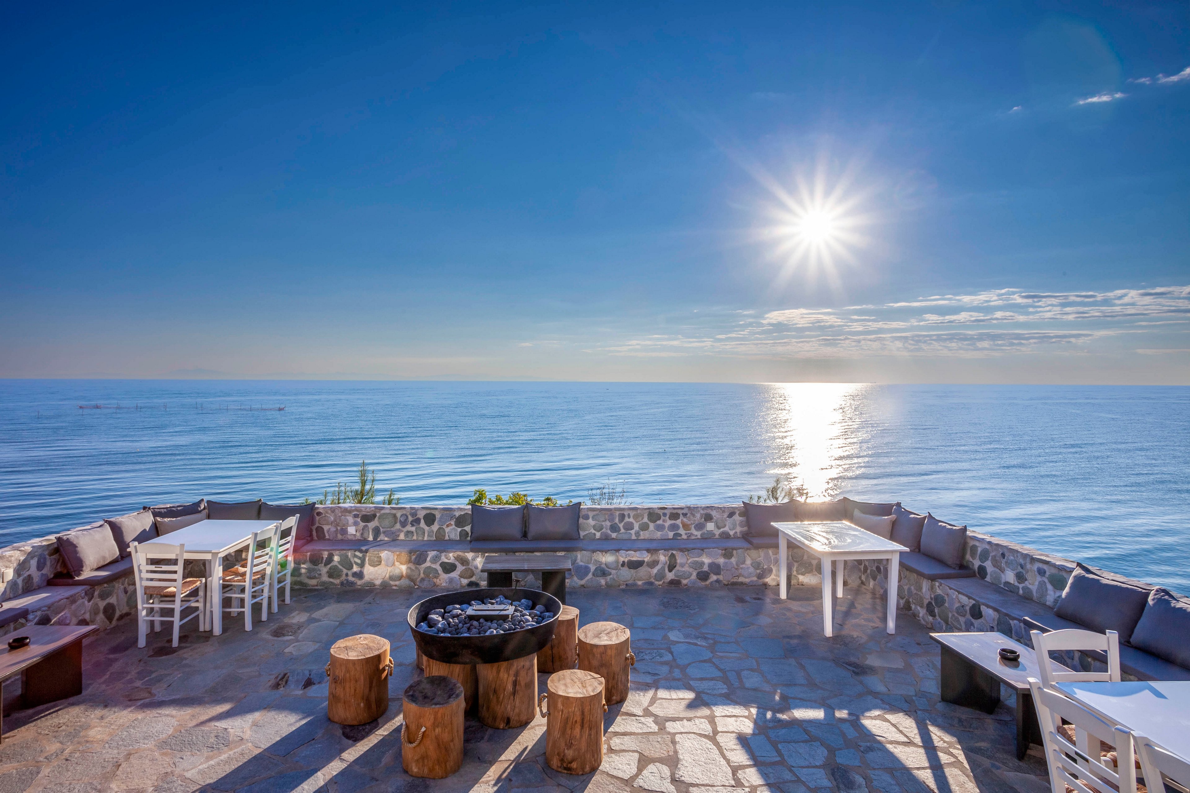 The main subject is a stylish outdoor seating area overlooking a calm ocean under a bright sun. The setting features stone flooring, wooden stools, white tables, and cushioned benches arranged around a central fire pit. The mood is tranquil and inviting, with a clear blue sky and sunlight reflecting off the water, creating a serene atmosphere. No visible text or numeric values are present in the image.