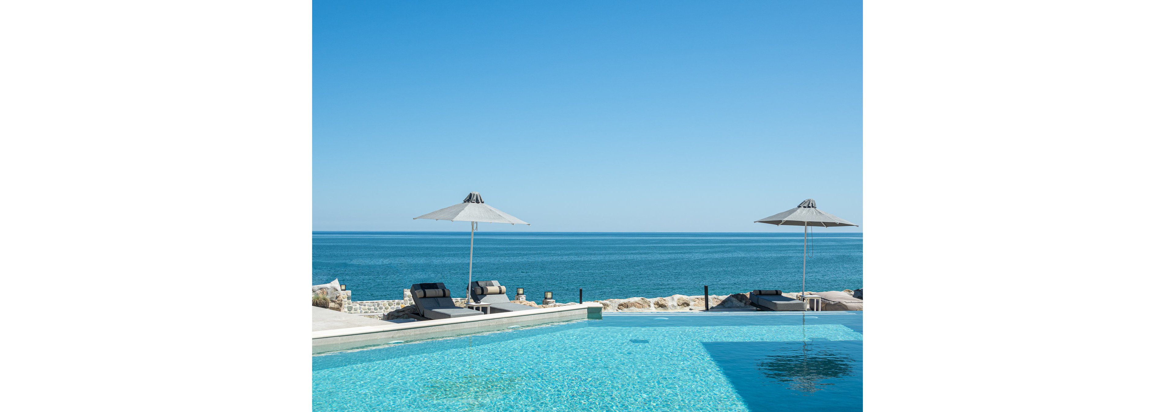A serene outdoor pool overlooks a calm blue ocean, with two modern lounge chairs and umbrellas providing shade. The setting is minimalist and tranquil, featuring clear skies and a sparkling turquoise pool. The scene evokes relaxation and leisure, with neutral tones and a clean, contemporary style. No visible text or numeric values are present in the image.